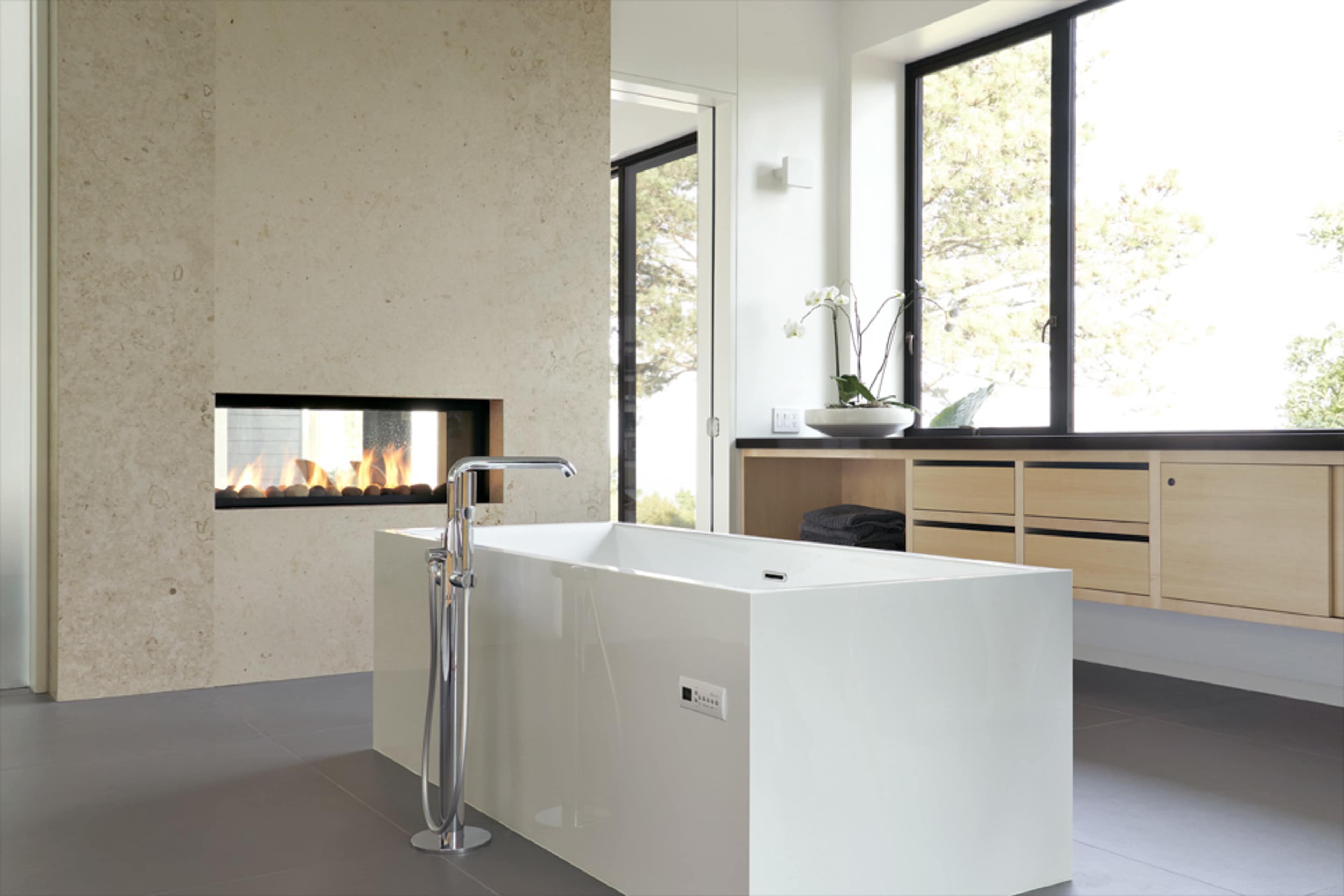 A modern bathroom featuring a freestanding white bathtub, a wall-mounted fireplace, large windows, and light wood cabinetry.
