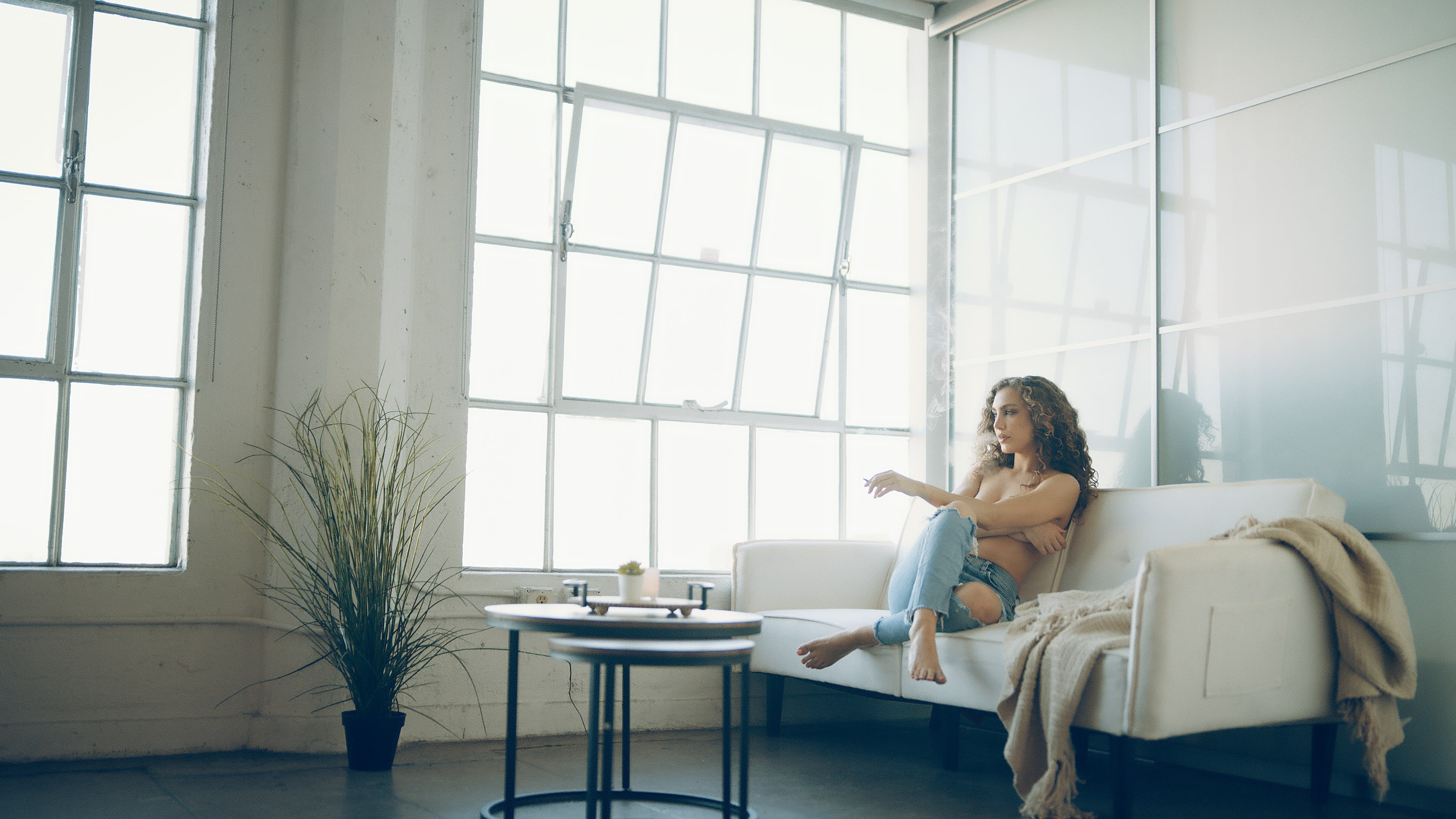 A woman sits on a white couch in a bright room with large windows, facing a small coffee table.