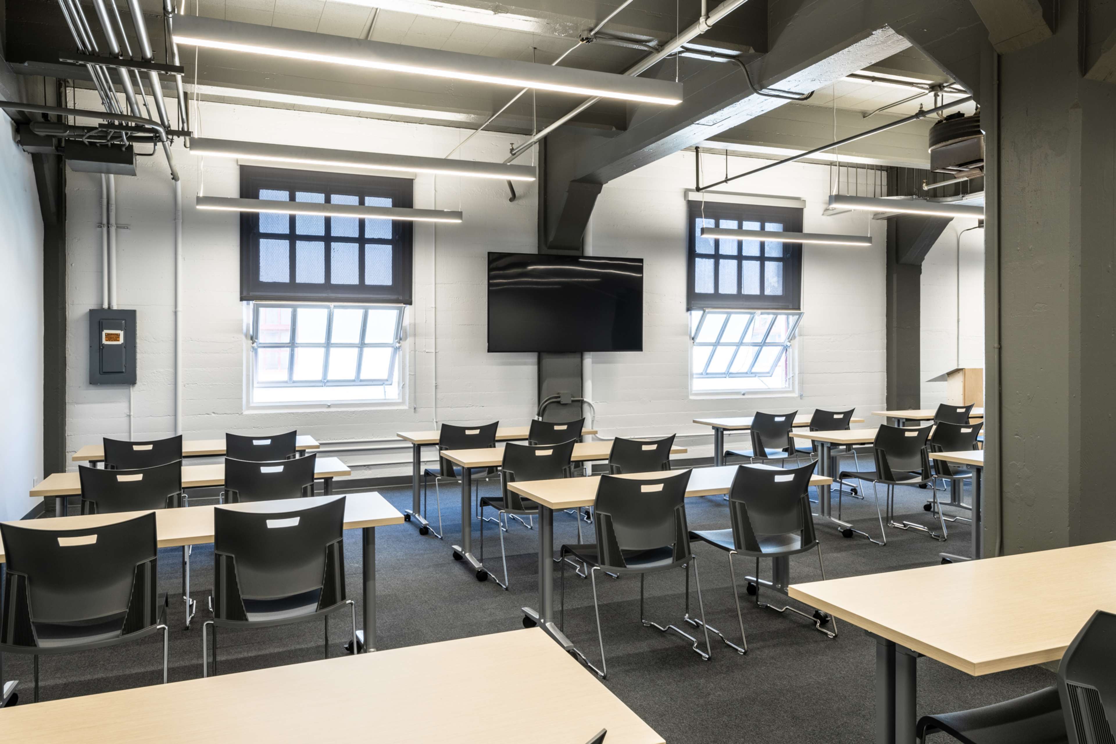 A classroom with rows of tables and chairs, a large screen on the wall, and windows providing natural light.