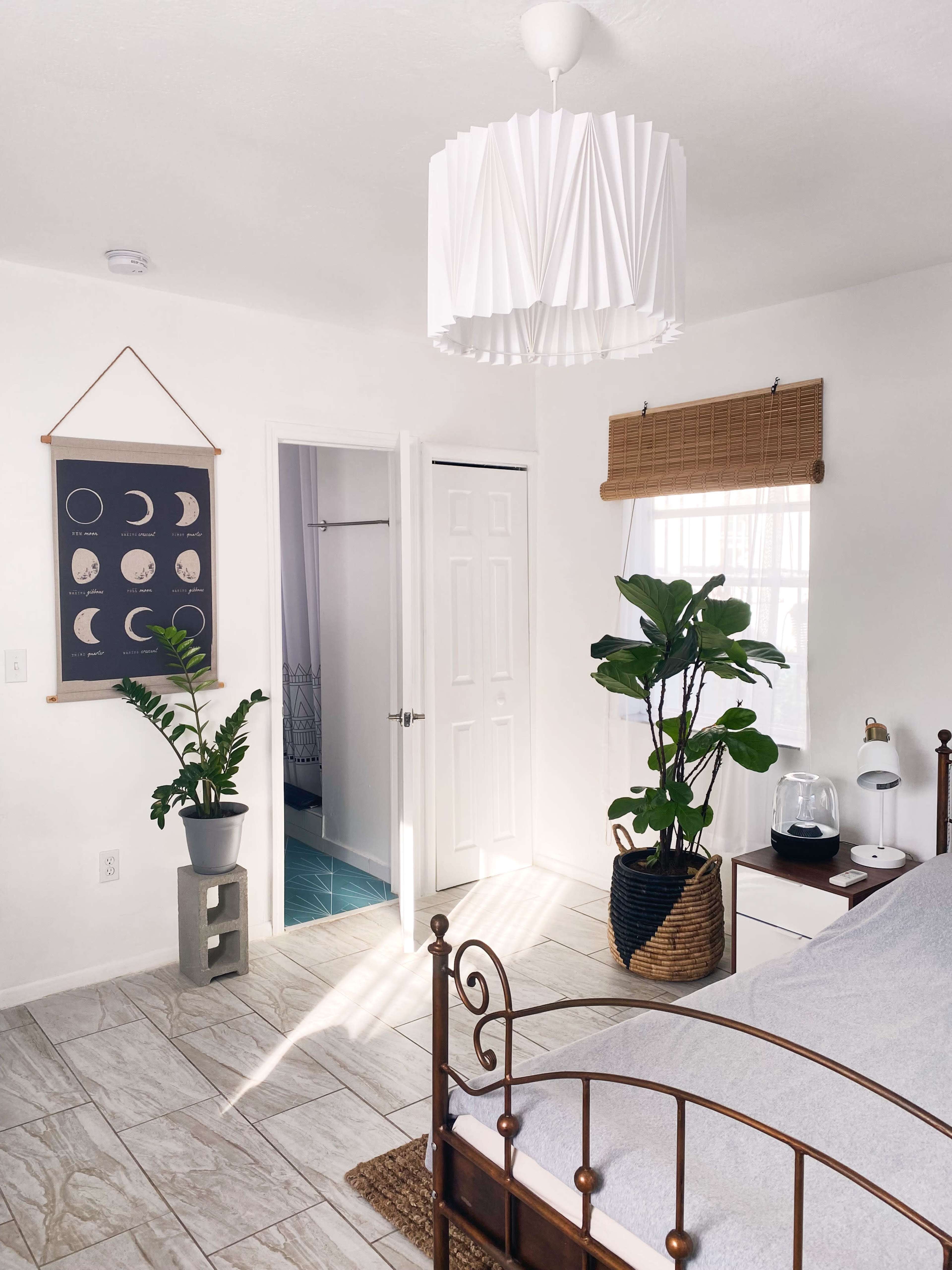 The room features a bed with a metal frame, a large pendant light, potted plants in the corners, and a door leading to a bathroom.