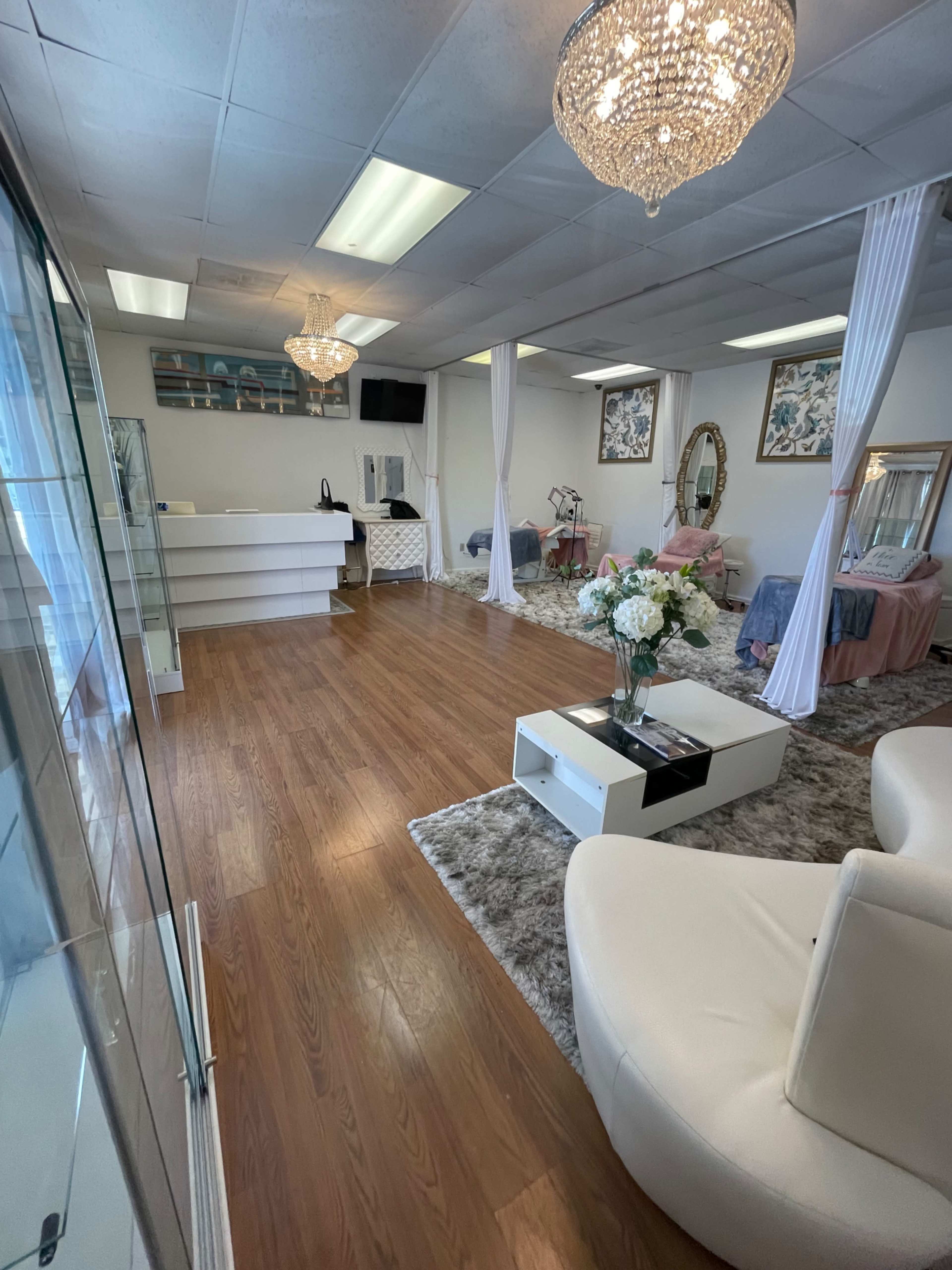 A spacious, well-lit salon interior featuring white furniture, decorative chandeliers, and treatment areas with draped curtains.