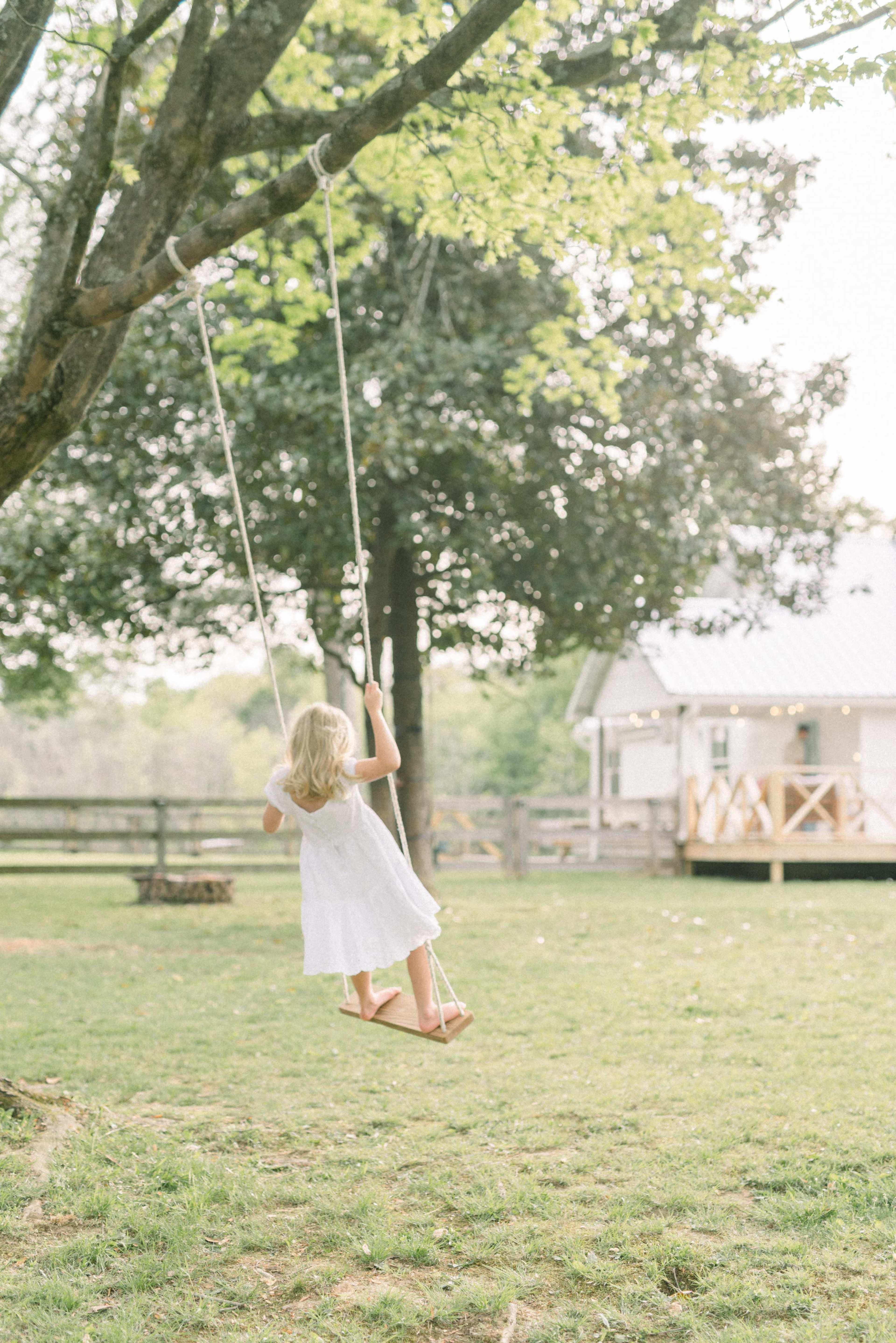 A young child in a white dress swings from a tree near a house in a grassy yard.