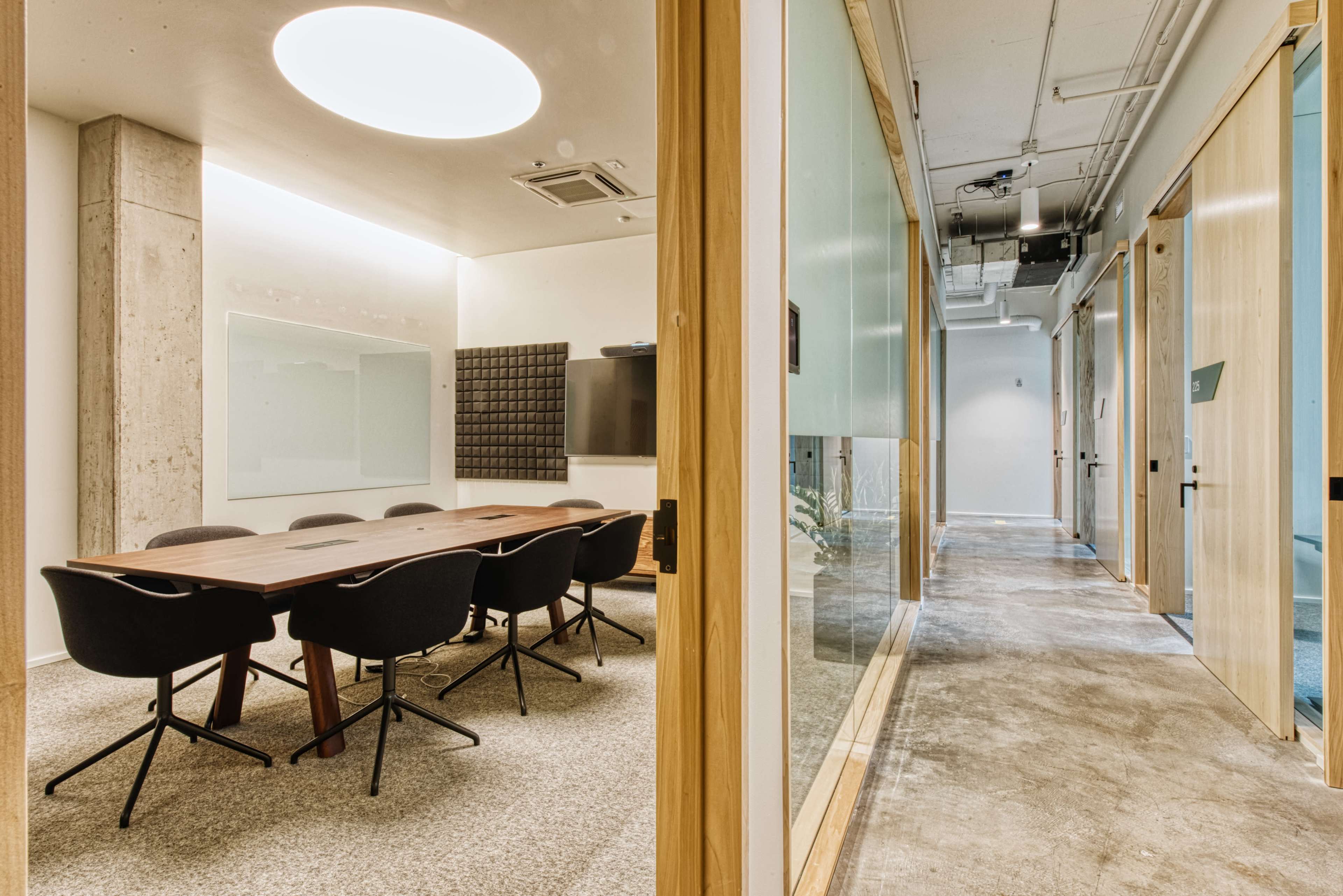 The image shows a modern conference room with a wooden table surrounded by black chairs, and a hallway leading to additional office spaces.