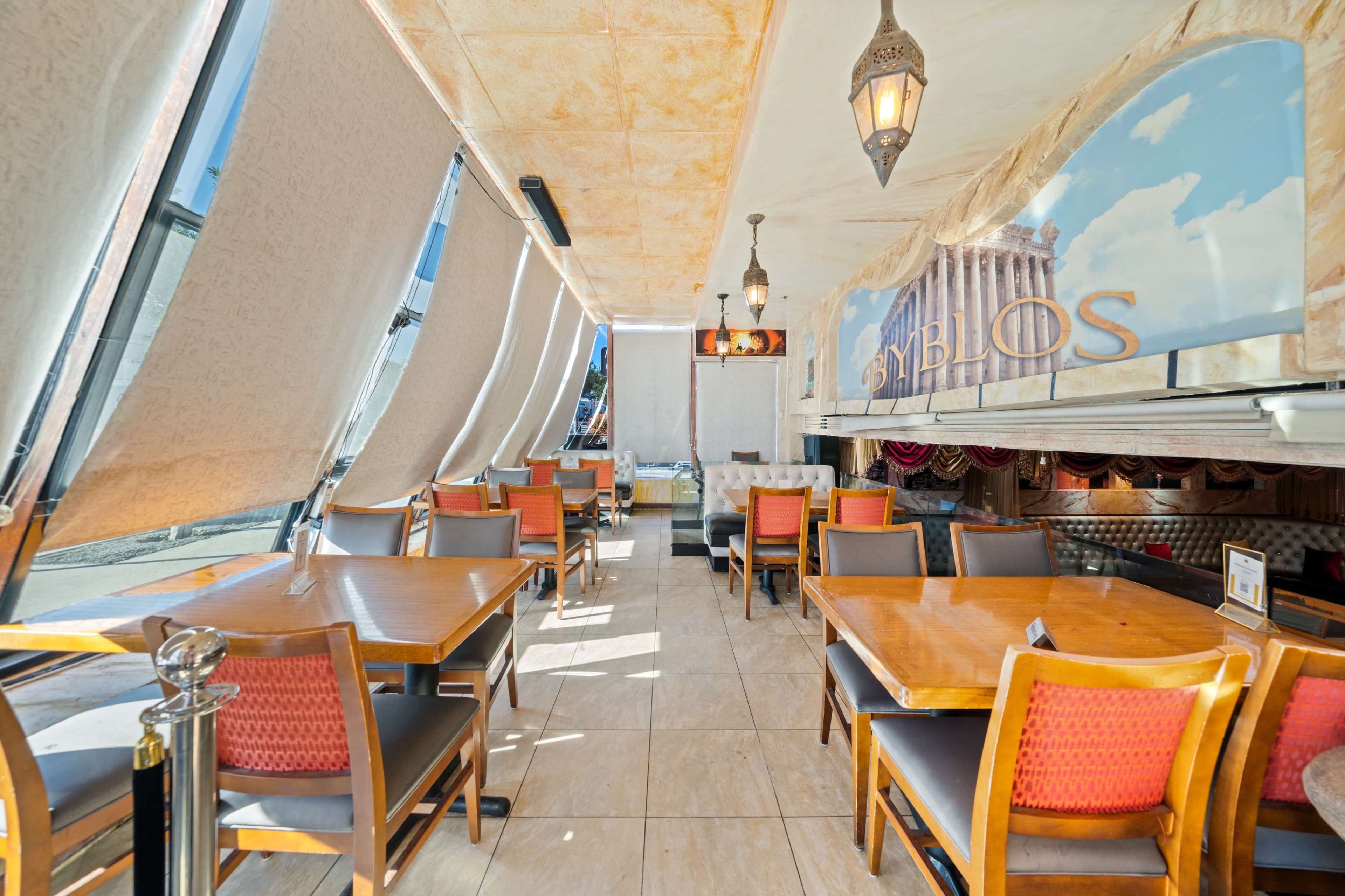 A restaurant interior with wooden tables and chairs, large windows with billowing shades, and classical-themed wall decor.