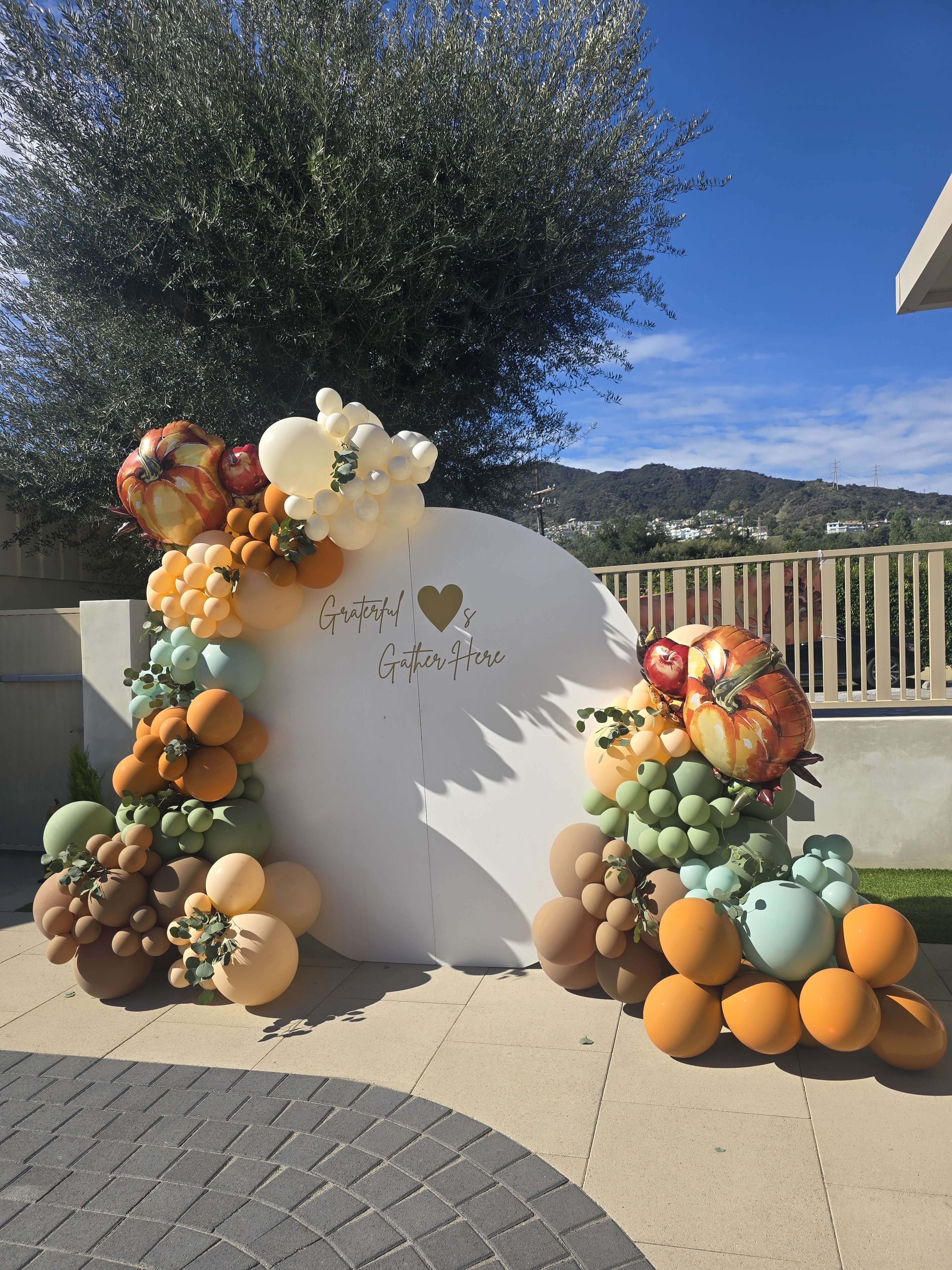 The image shows a decorative balloon arch composed of various colored balloons, including orange, green, and brown, alongside a white circular backdrop with the words "Grateful" and "Gather Here."