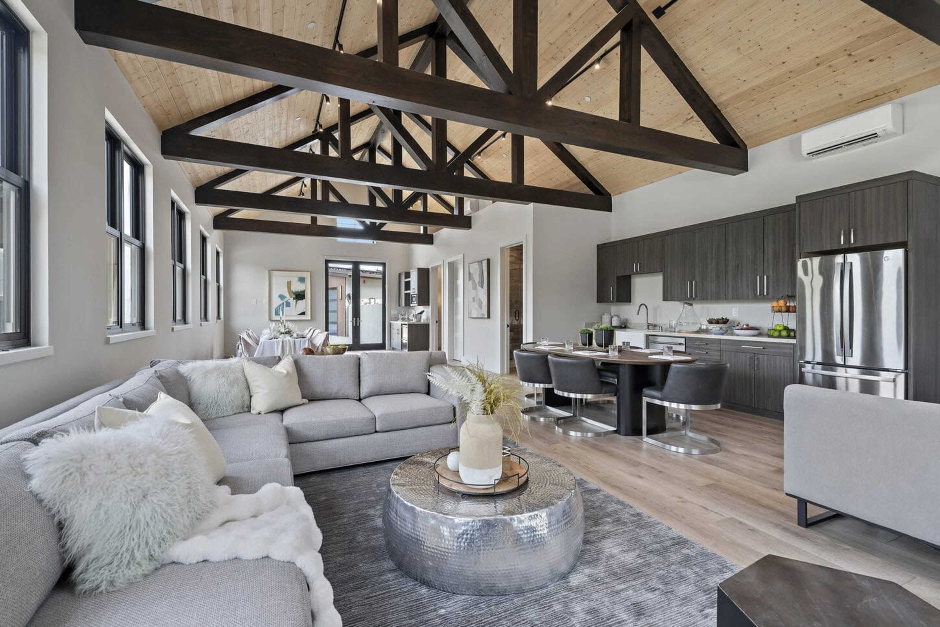 The image shows a modern open-concept living space with a gray sectional sofa, wooden beams in the ceiling, and a kitchen area featuring dark cabinetry and stainless steel appliances.