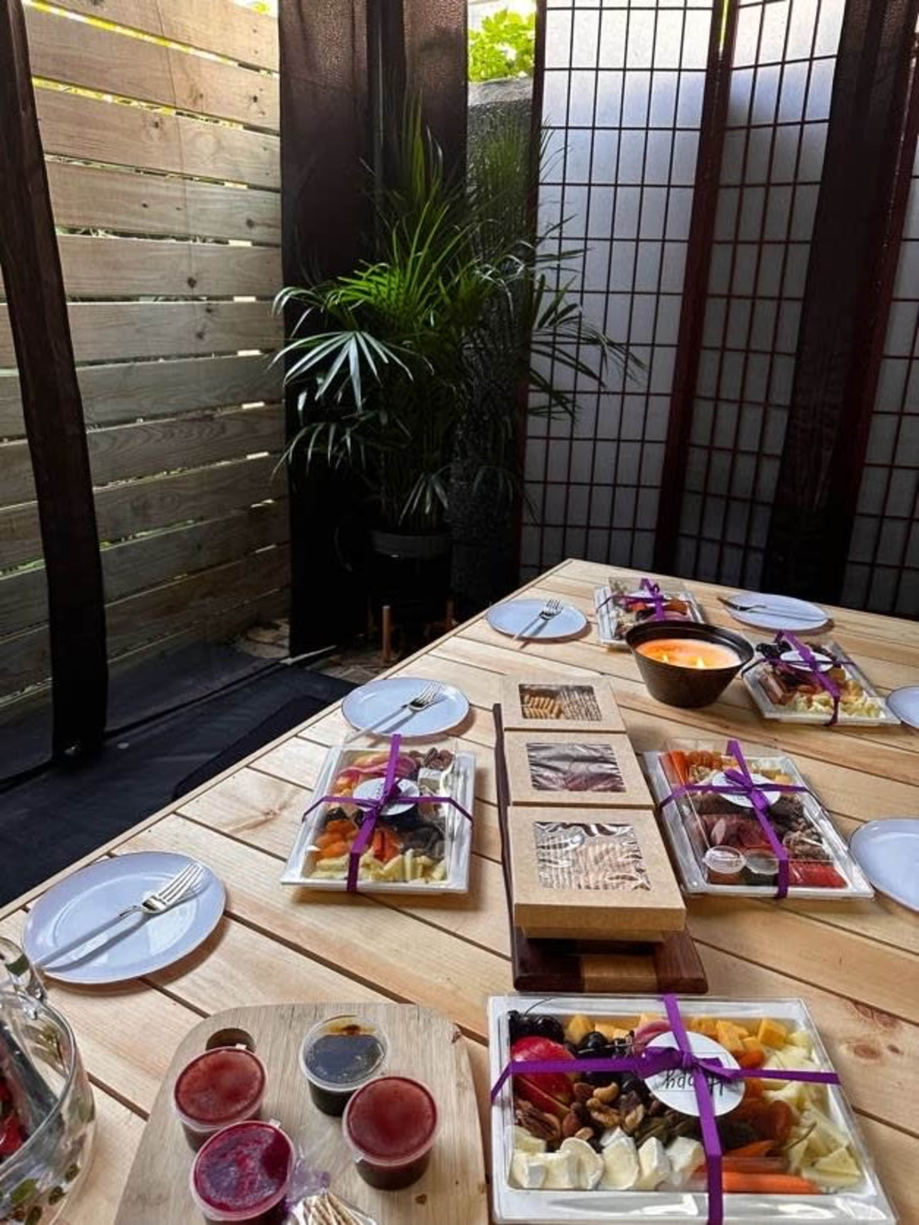 A wooden table is set outdoors with multiple plates of food, sauces, and a bowl, surrounded by partitions and greenery.