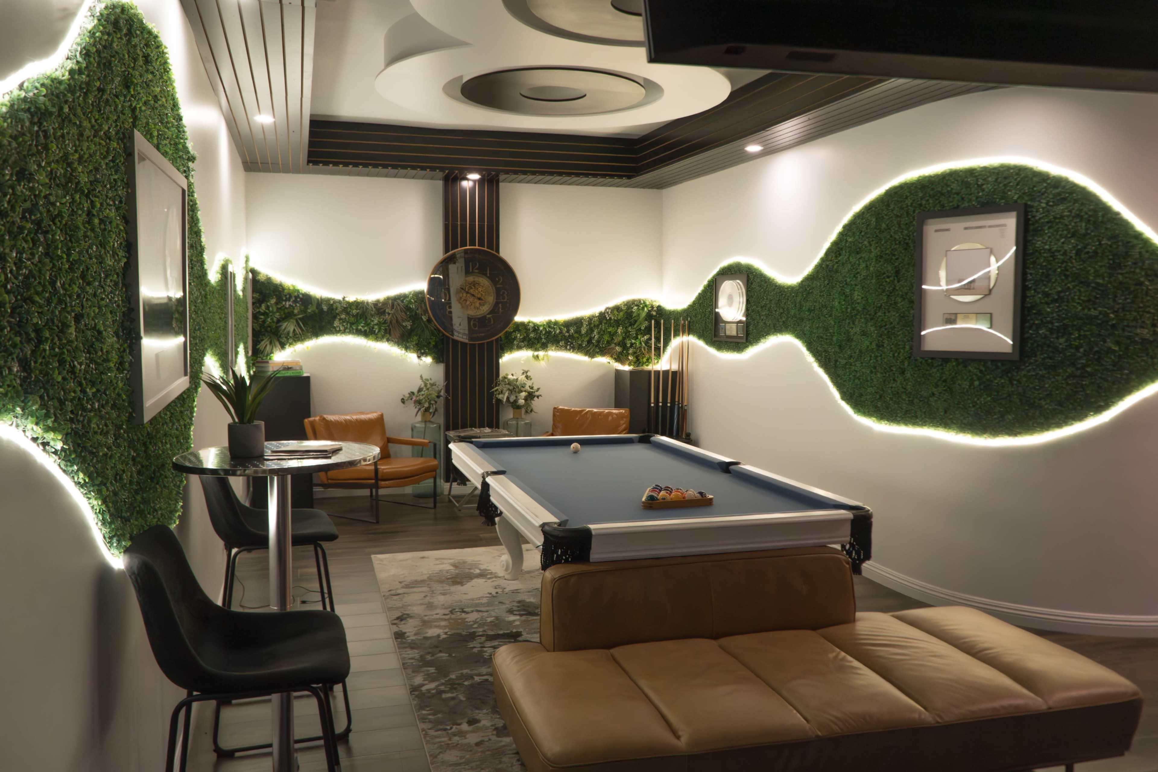 The image shows a modern recreational room featuring a pool table, a lounge area with a sofa, and decorative wall elements covered in greenery.