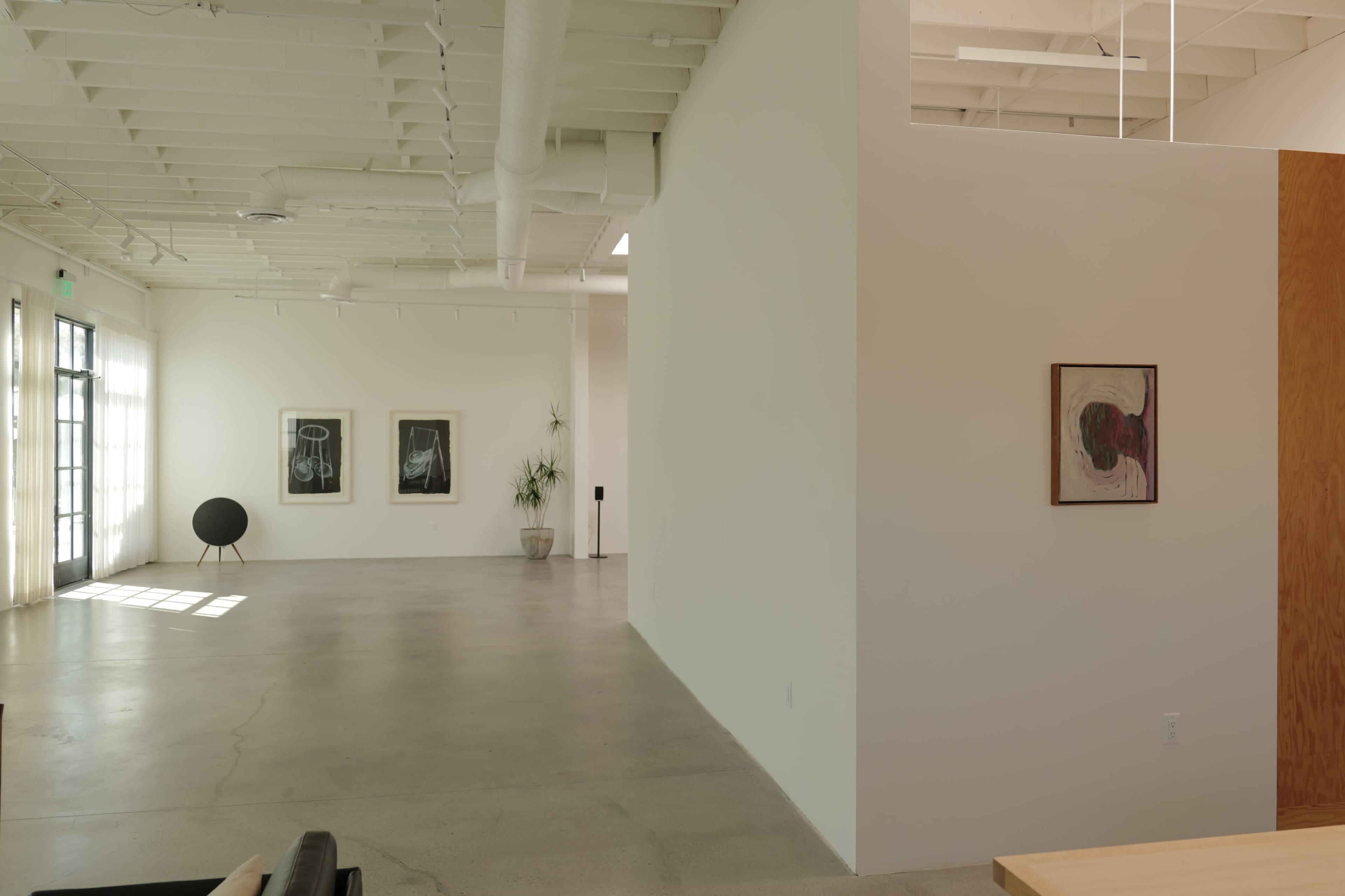 The image shows a modern art gallery featuring minimalistic design, with white walls, polished concrete flooring, and several framed artworks displayed on the walls.