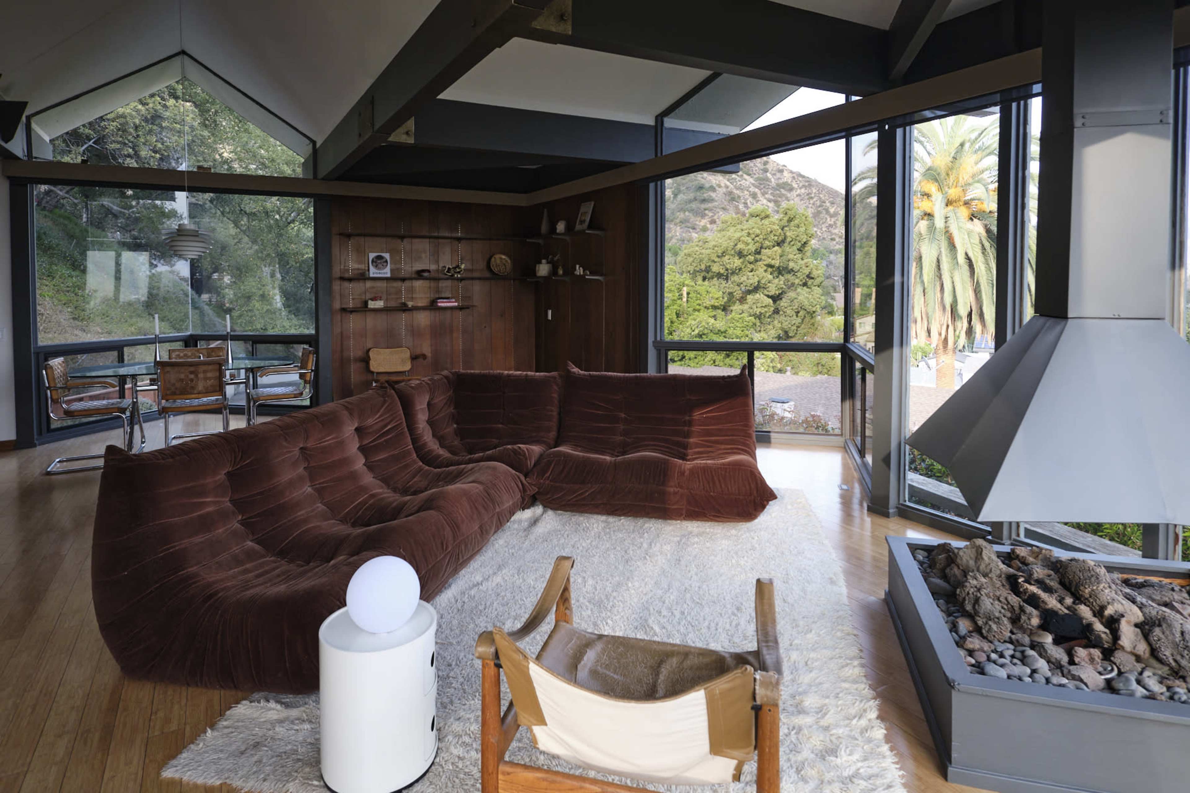 A modern living room with large windows, featuring a brown sectional sofa, a light-colored rug, a fireplace, and wooden accents.