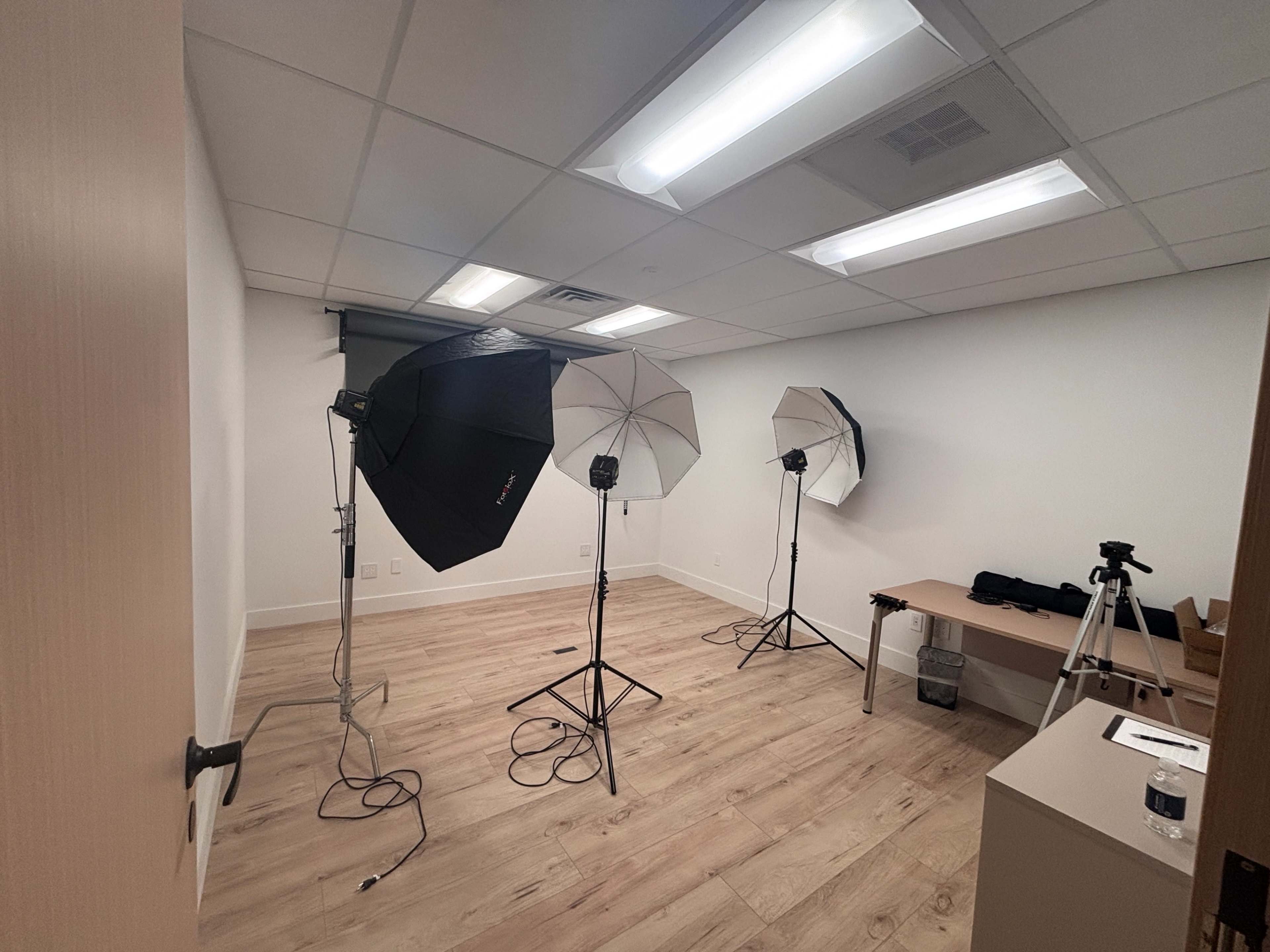 The image shows a well-lit studio room with three studio light setups on stands and a table against one wall.