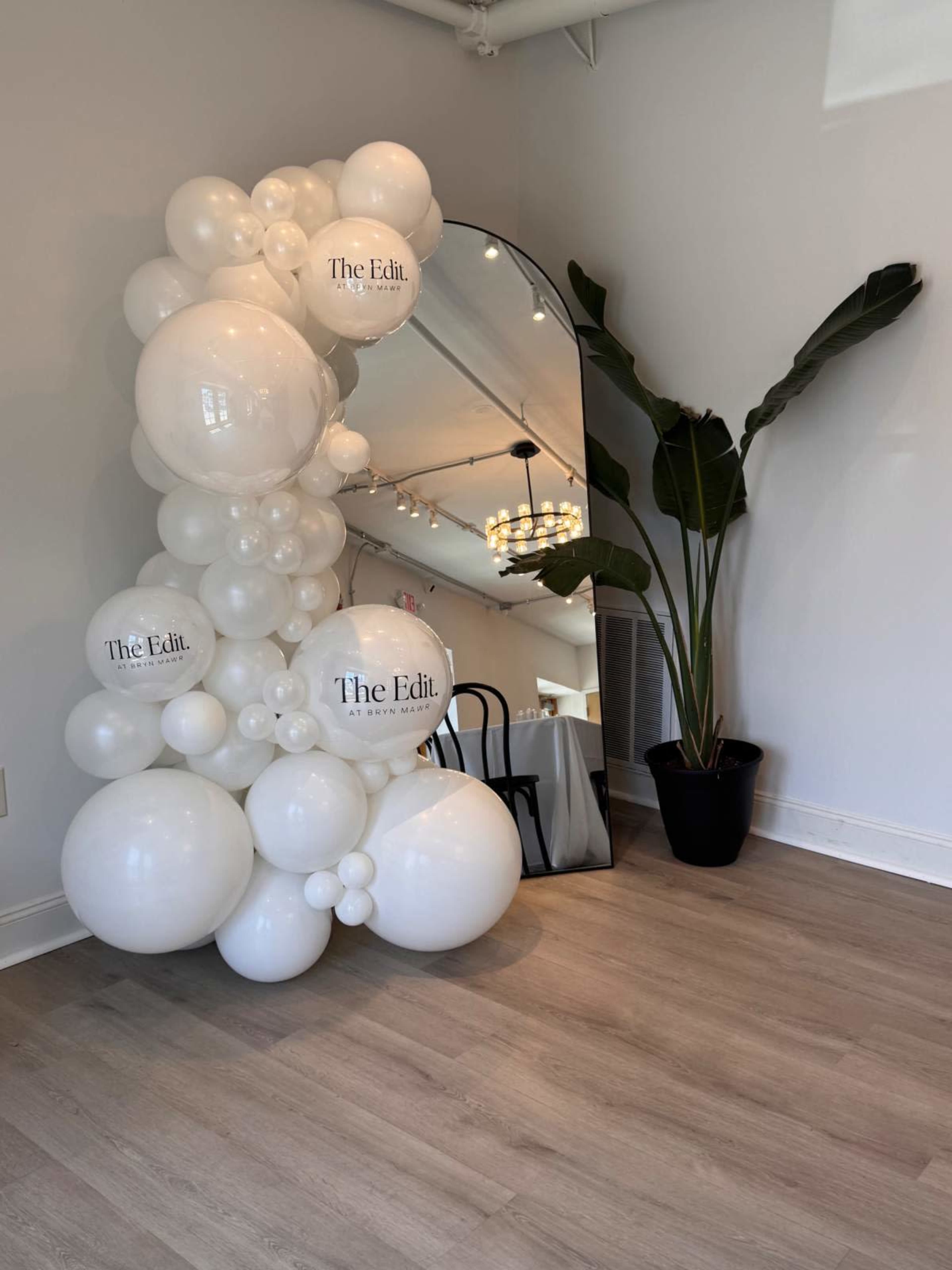 The image shows a decorative arch made of white balloons with the text "The Edit." beside a tall plant and a large mirror in a light-colored room.
