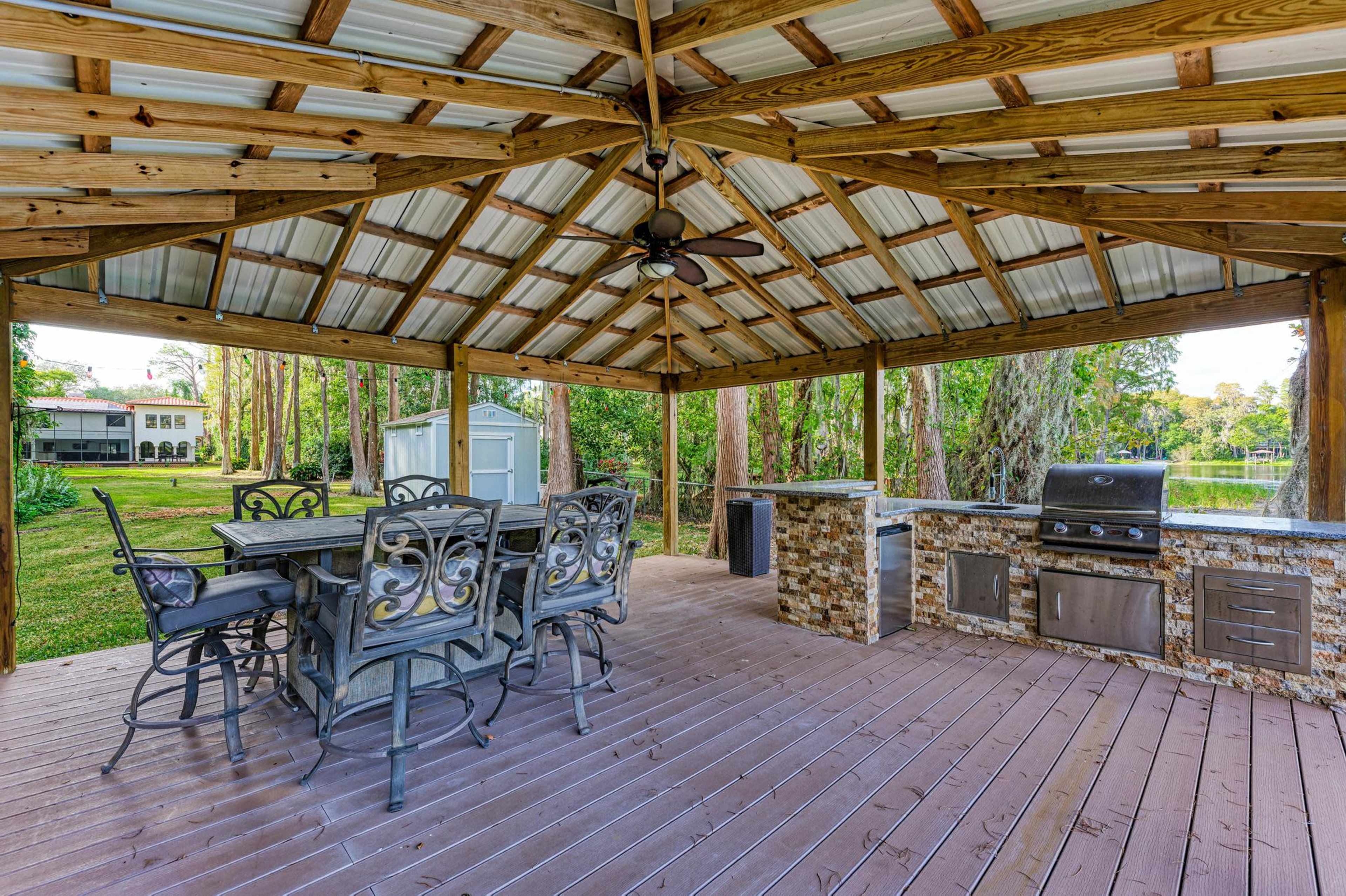 A covered outdoor pavilion with a dining area featuring a table and chairs, alongside a built-in kitchen space with a grill, set in a landscaped yard.