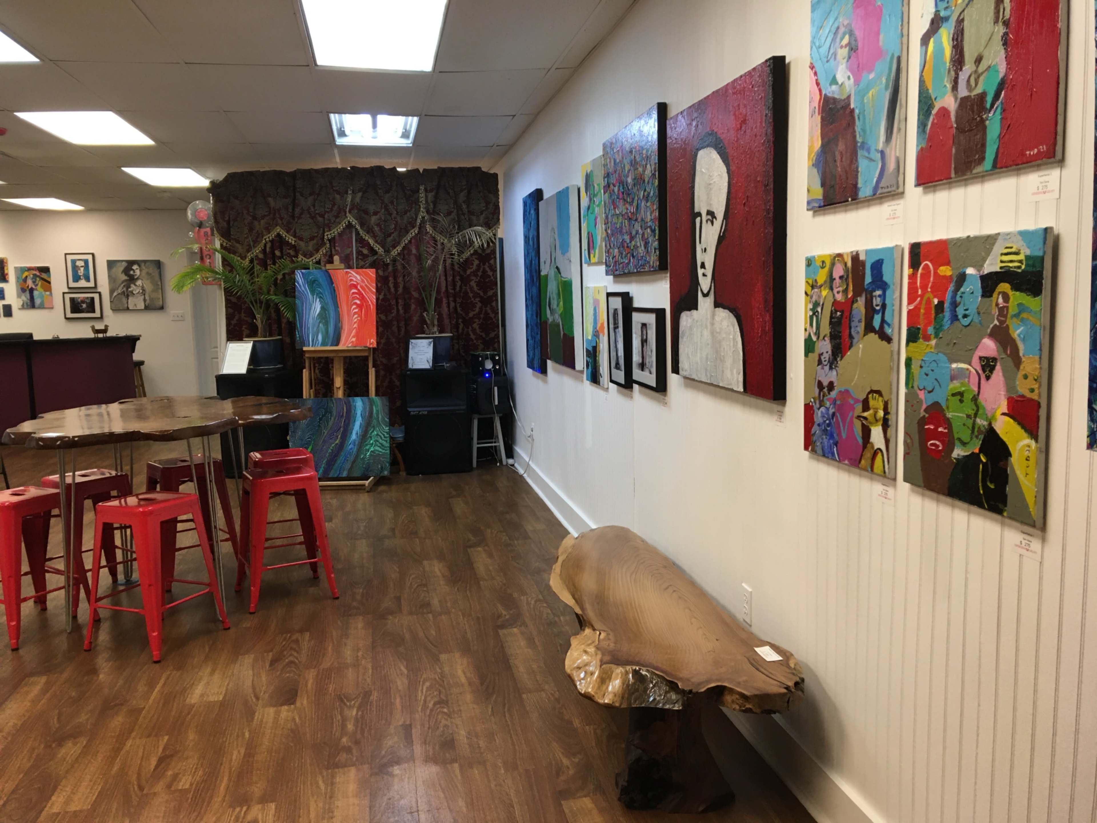 The image shows an art gallery featuring colorful paintings on the walls, a wooden table with red stools, and a wooden bench in the foreground.