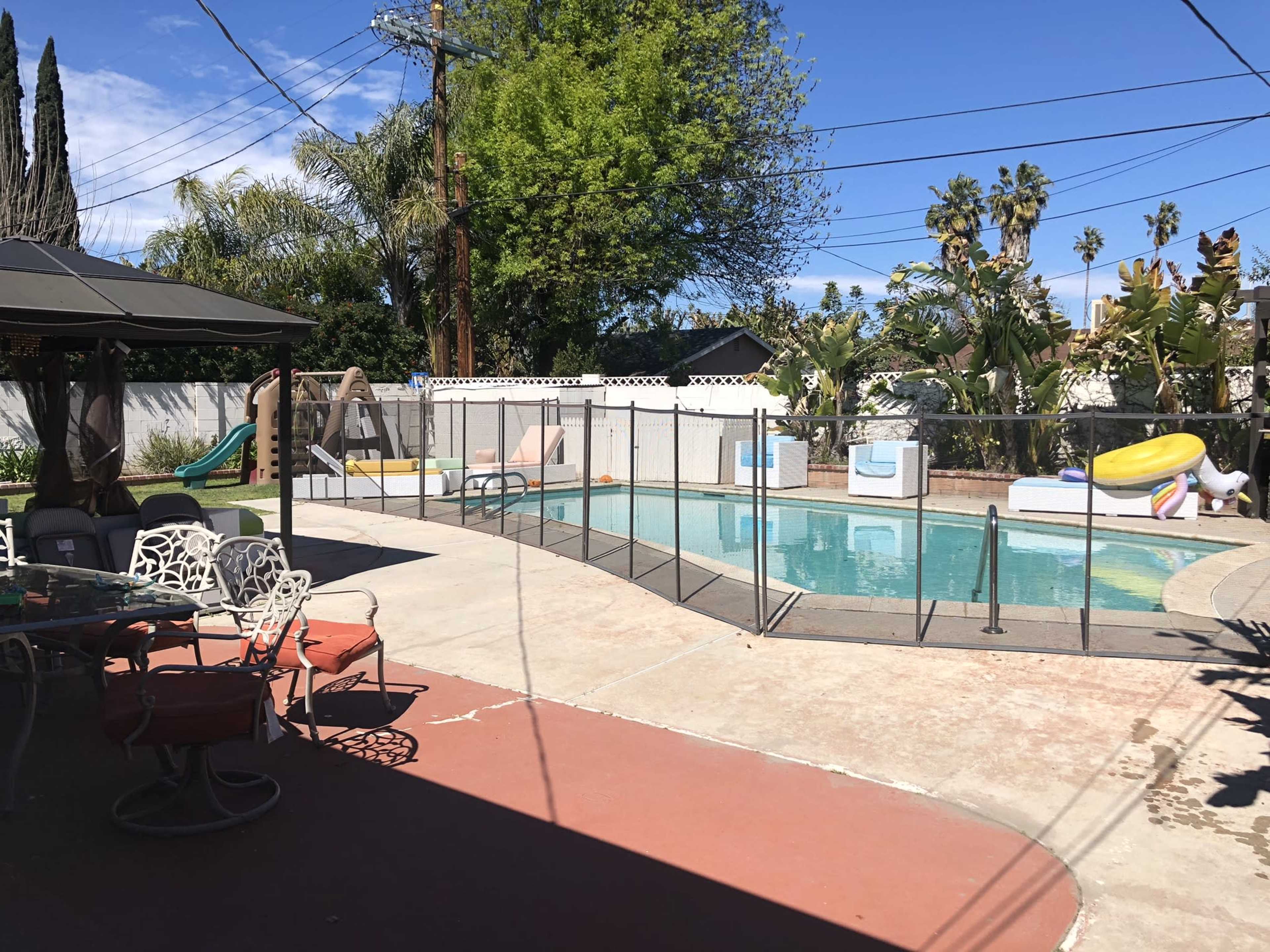 A residential backyard featuring a swimming pool surrounded by a safety fence, lounge chairs, and a playground set in a sunny environment.