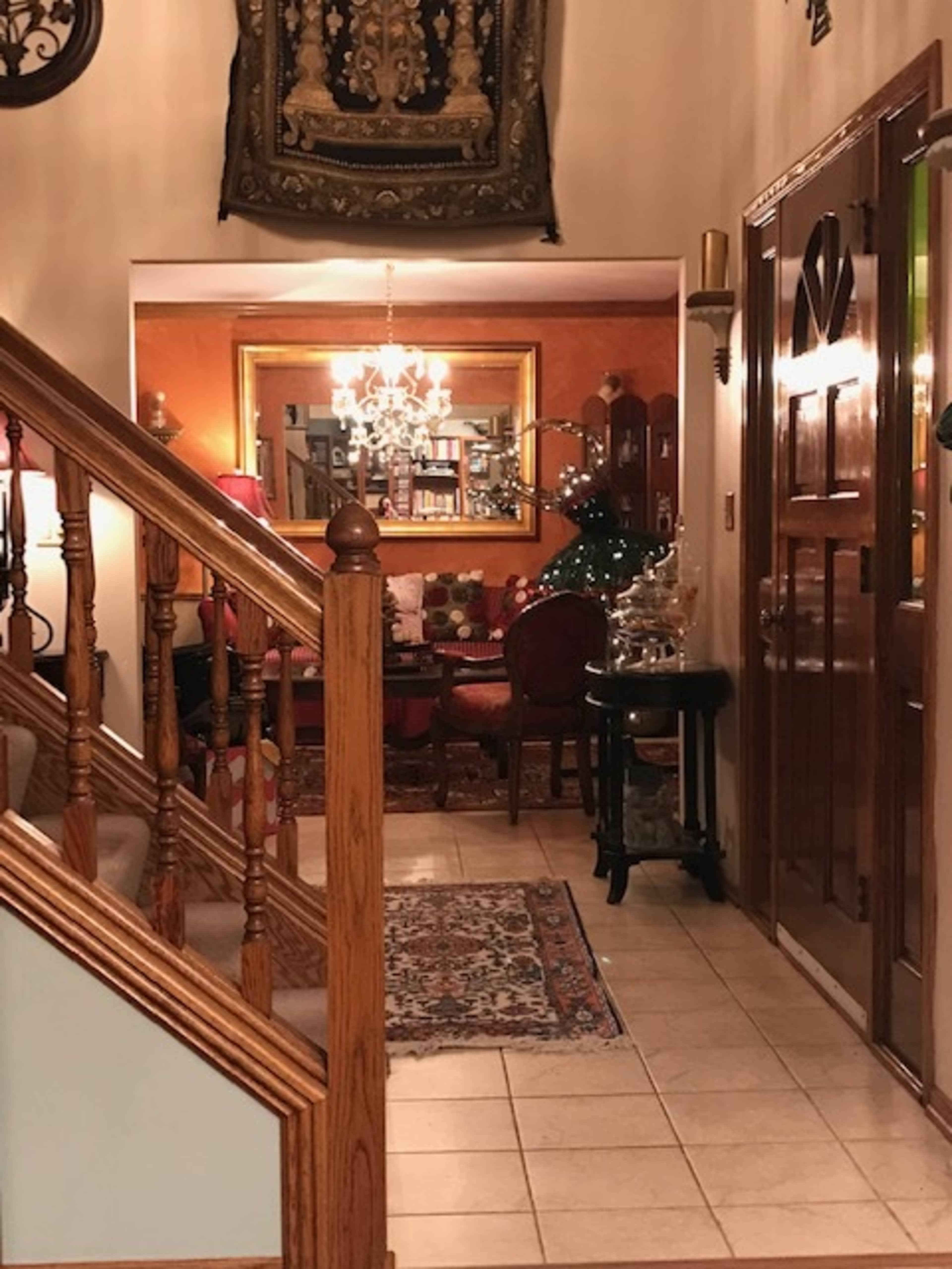 The image shows a warmly lit entryway with a wooden staircase, a decorative rug on tiled flooring, and a cozy sitting area visible in the background.