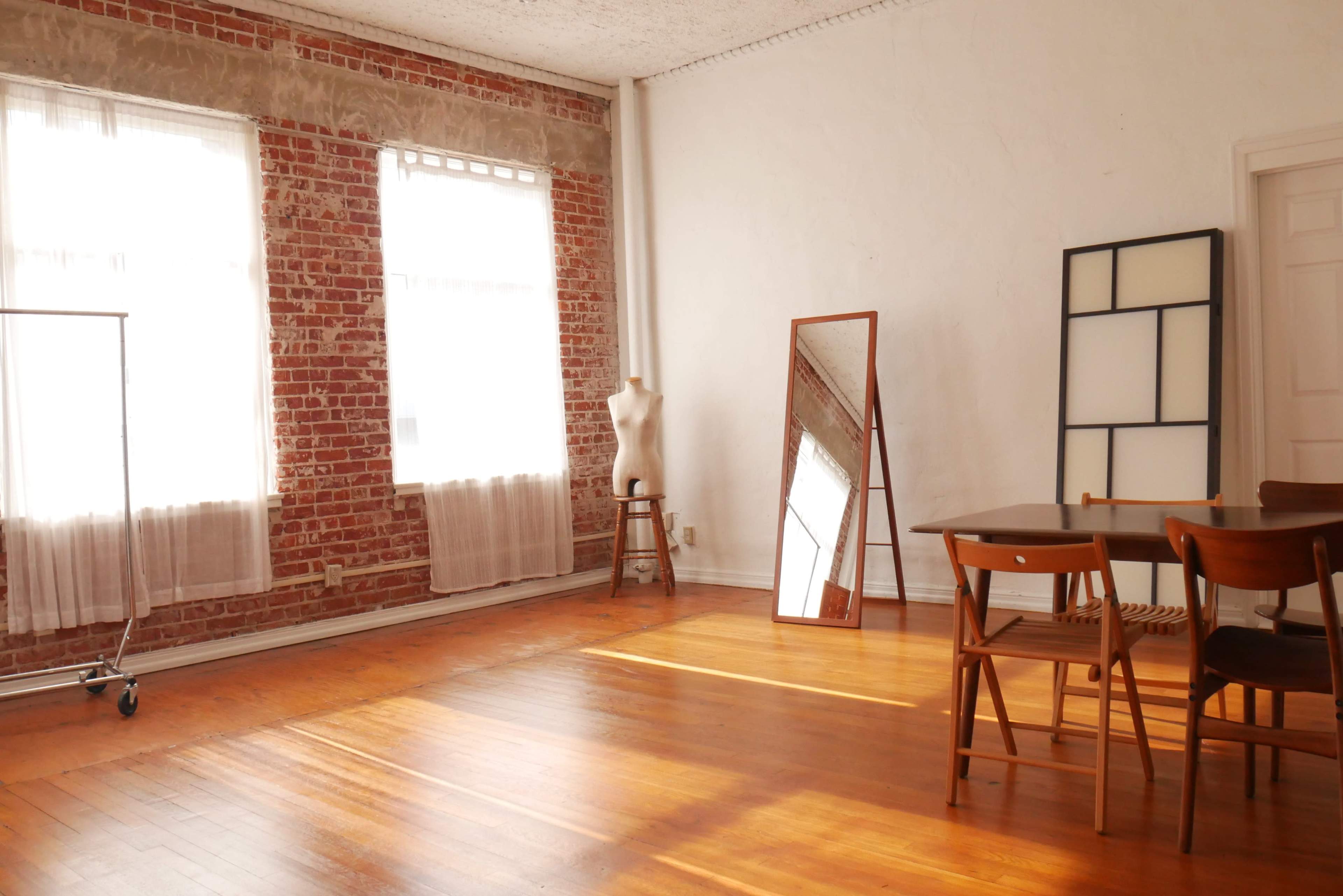 A spacious room with brick walls, large windows letting in light, a wooden floor, a mirror, a mannequin on a stool, and a dining table with chairs.