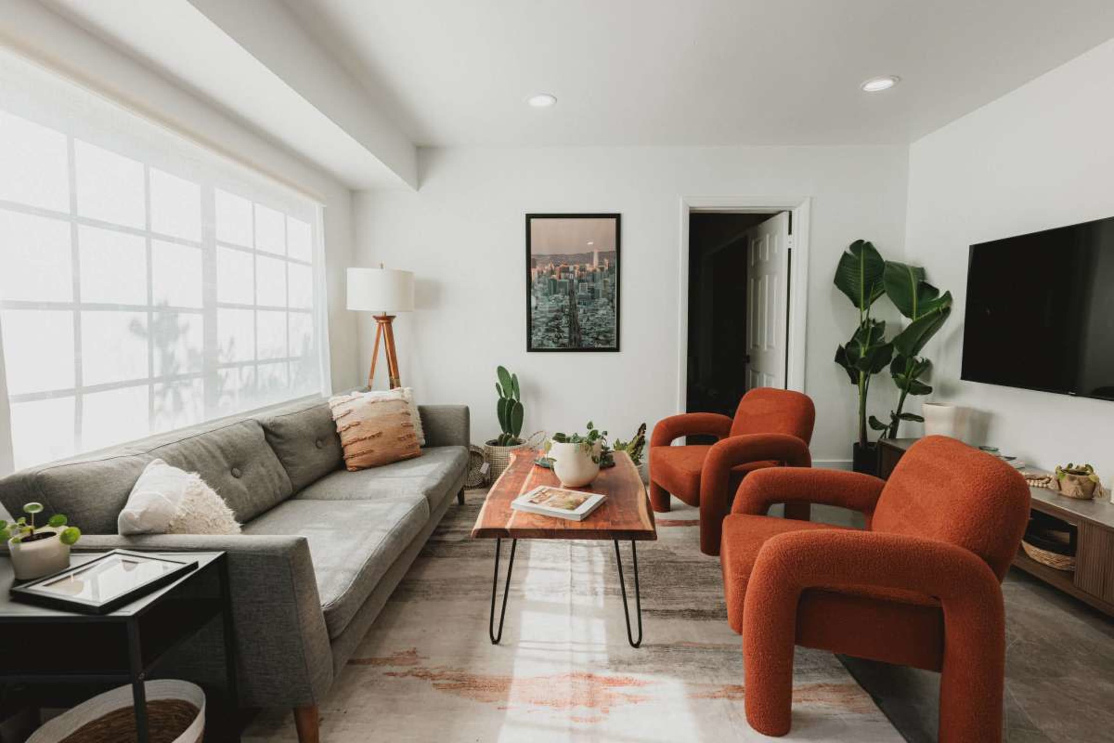 The image depicts a modern living room featuring a gray sofa, two orange chairs, a wooden coffee table, and various plants, with a large window allowing natural light to enter.