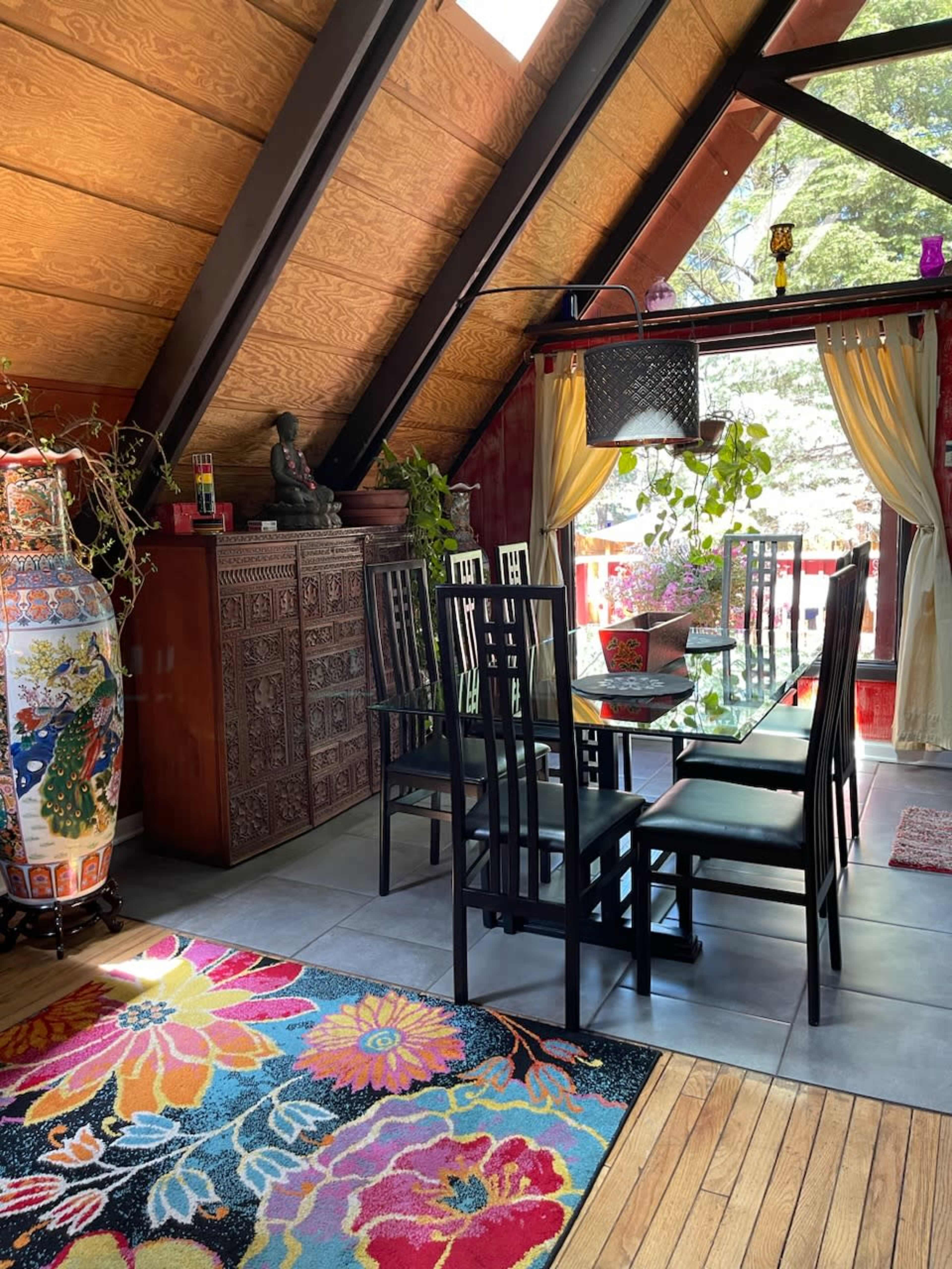 The interior features a dining area with a glass table surrounded by black chairs, wooden beams, and colorful decor, including plants and art pieces.