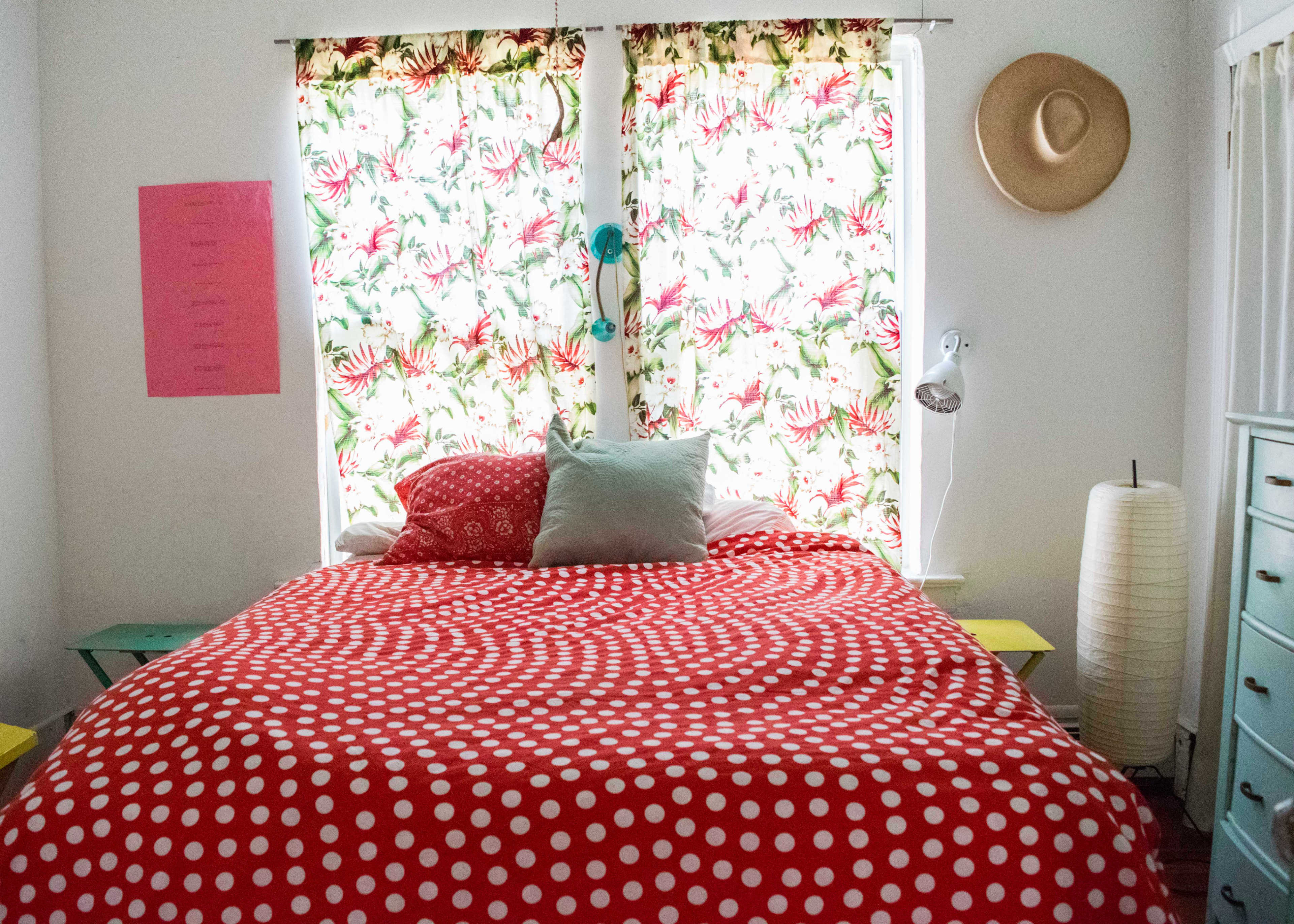 A bedroom with a large bed covered in a red polka dot comforter, surrounded by floral curtains, a bedside lamp, and a hat hanging on the wall.