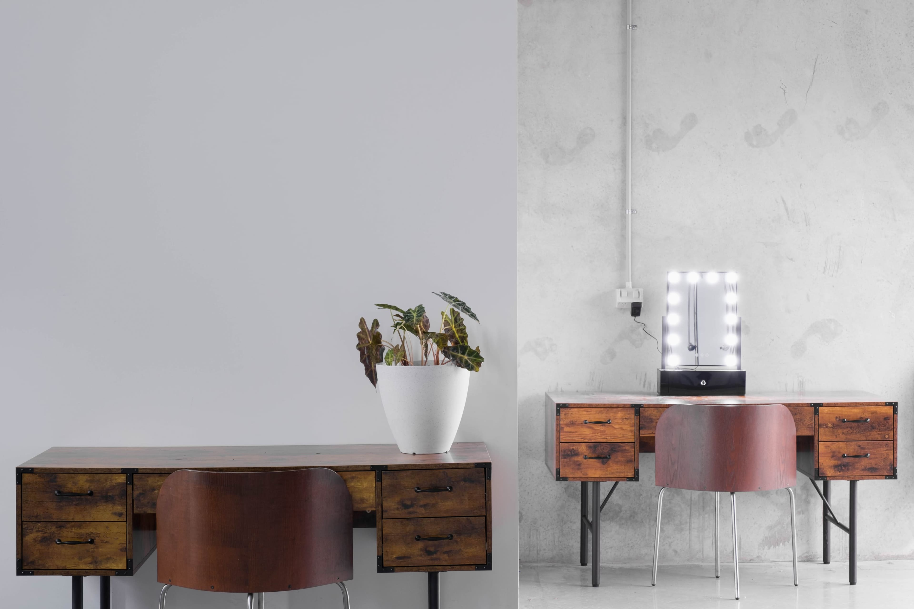 Two contrasting desks: one made of wooden drawers with a plant in a white pot on the left, and a sleek, modern desk with a lighted mirror on the right against a concrete wall.