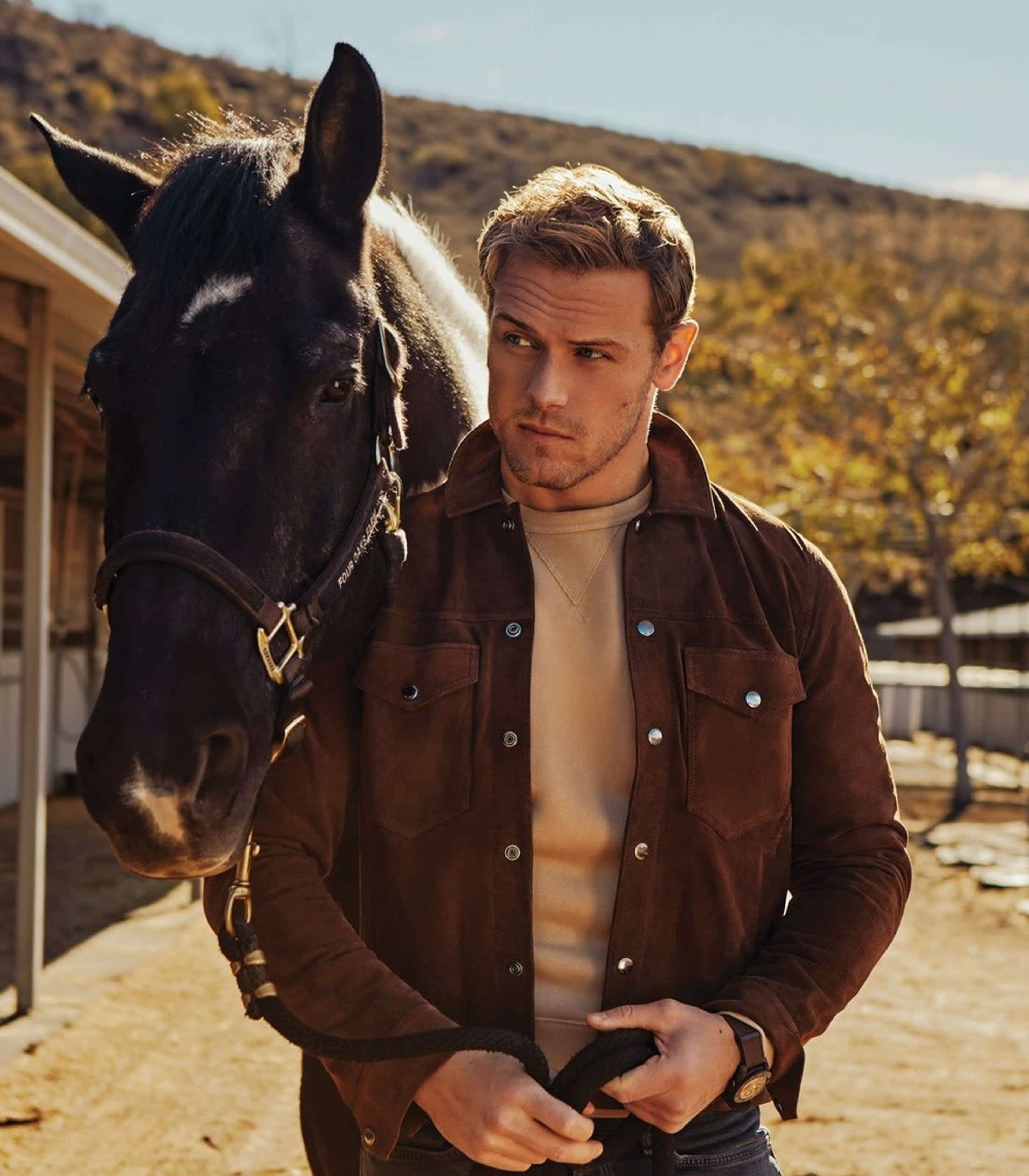 A young man in a brown suede jacket stands next to a black horse in a stable area with a sunlit background.