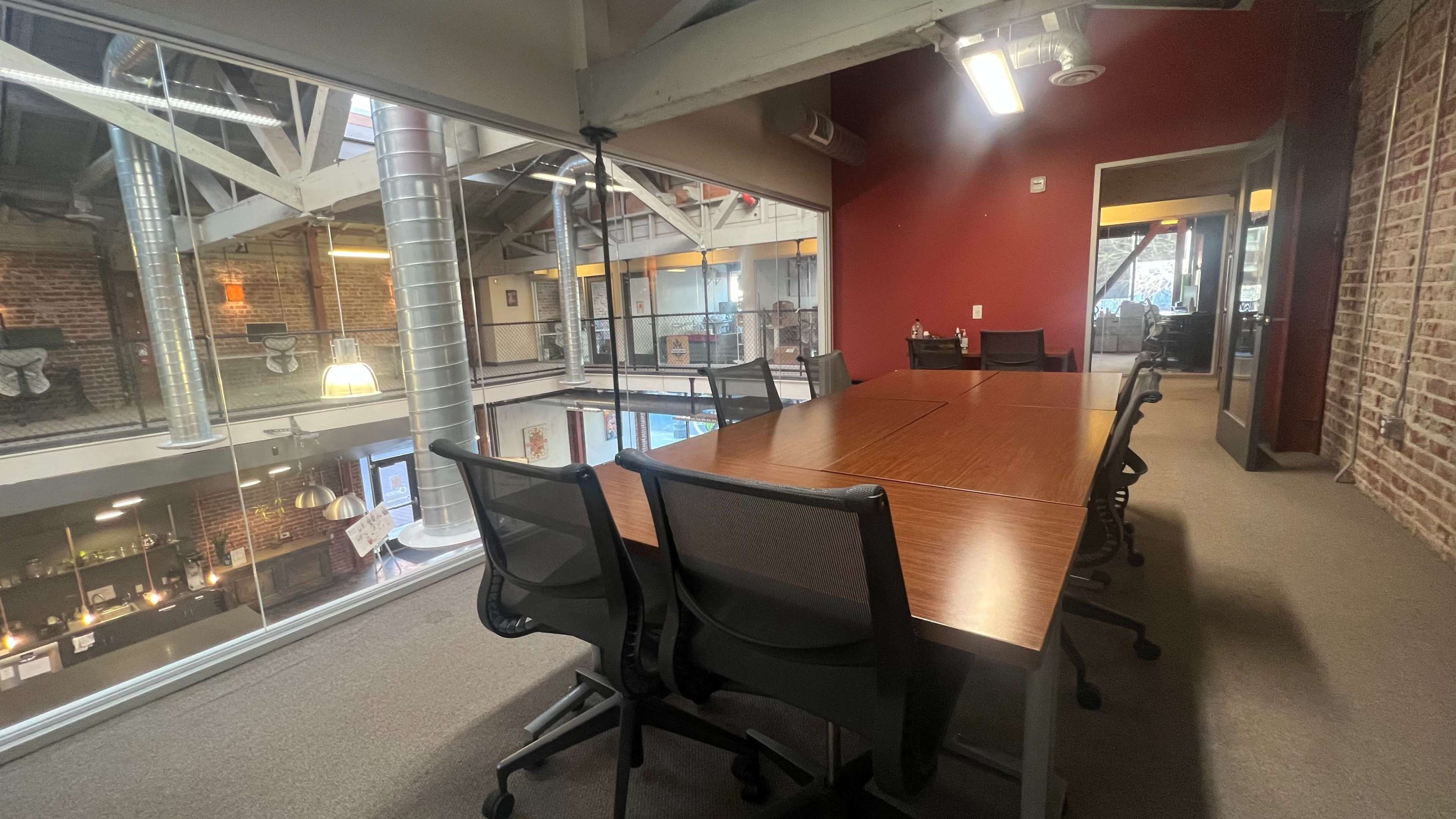 A large conference table is set with black chairs in a modern office space featuring exposed brick walls and large windows.