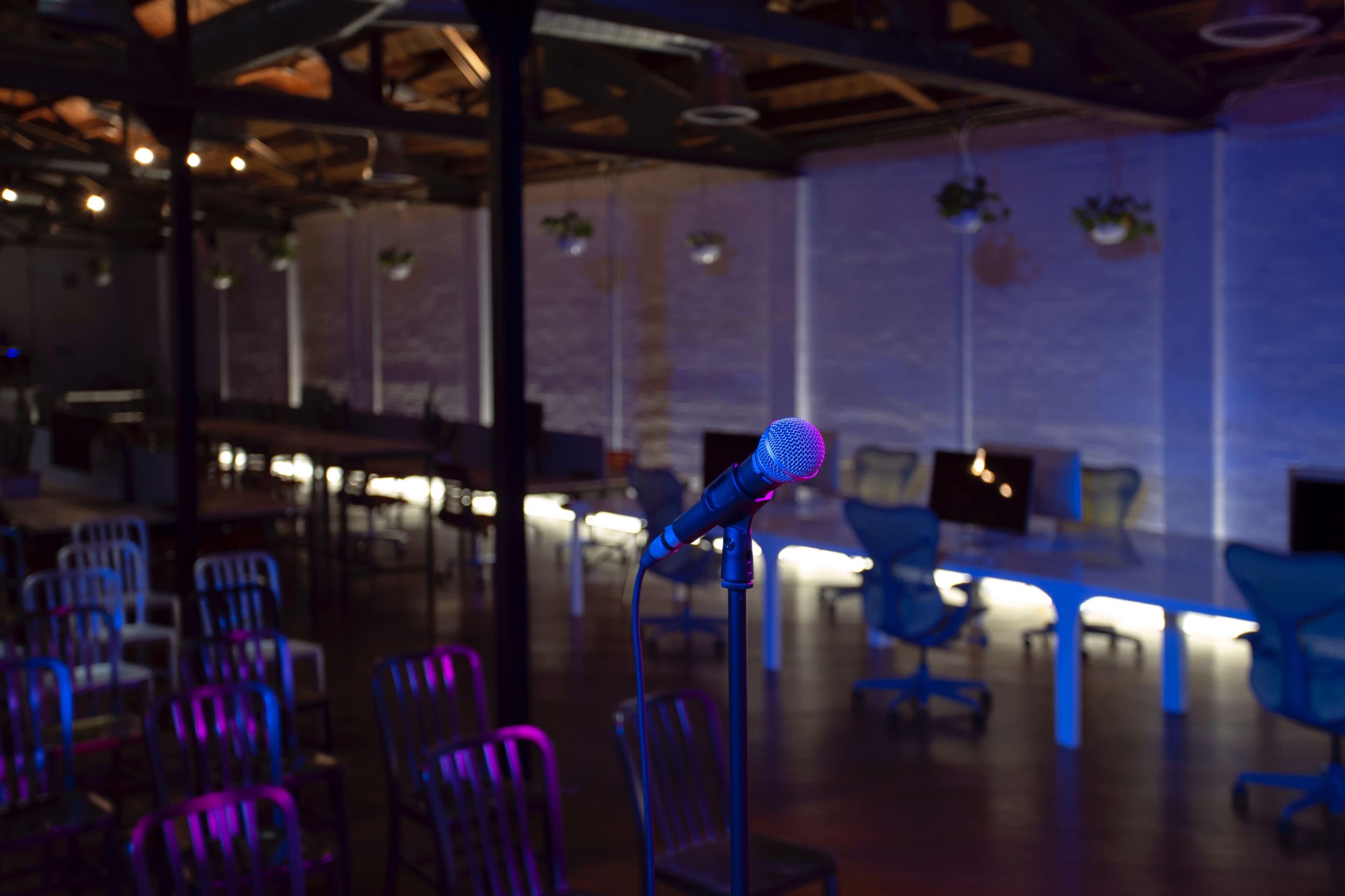 A microphone stands in the foreground of an empty event space with rows of chairs and illuminated workstations in the background.