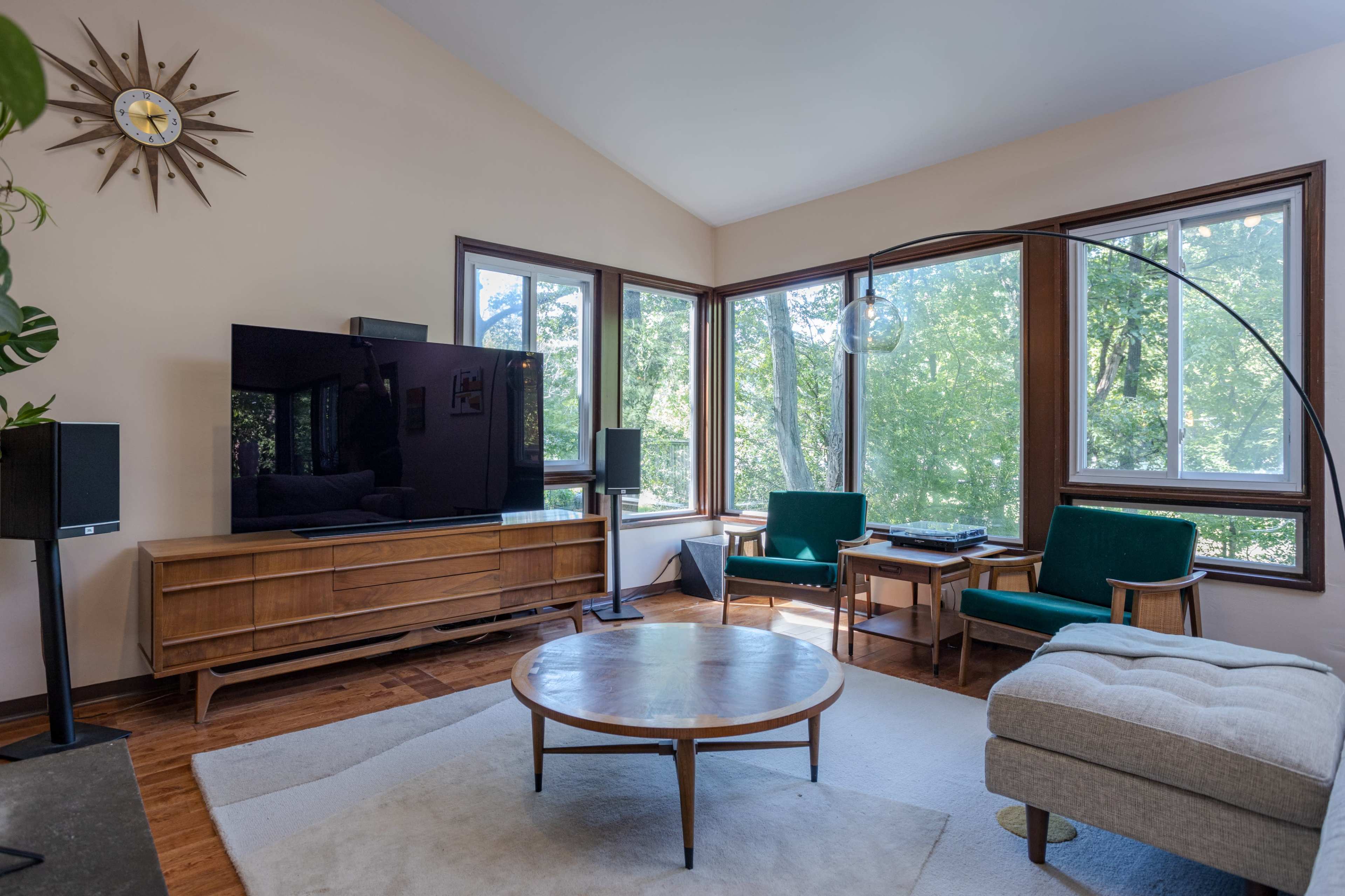 A modern living room features a large television on a wooden cabinet, surrounded by green chairs and a circular coffee table, with large windows providing a view of the outdoors.