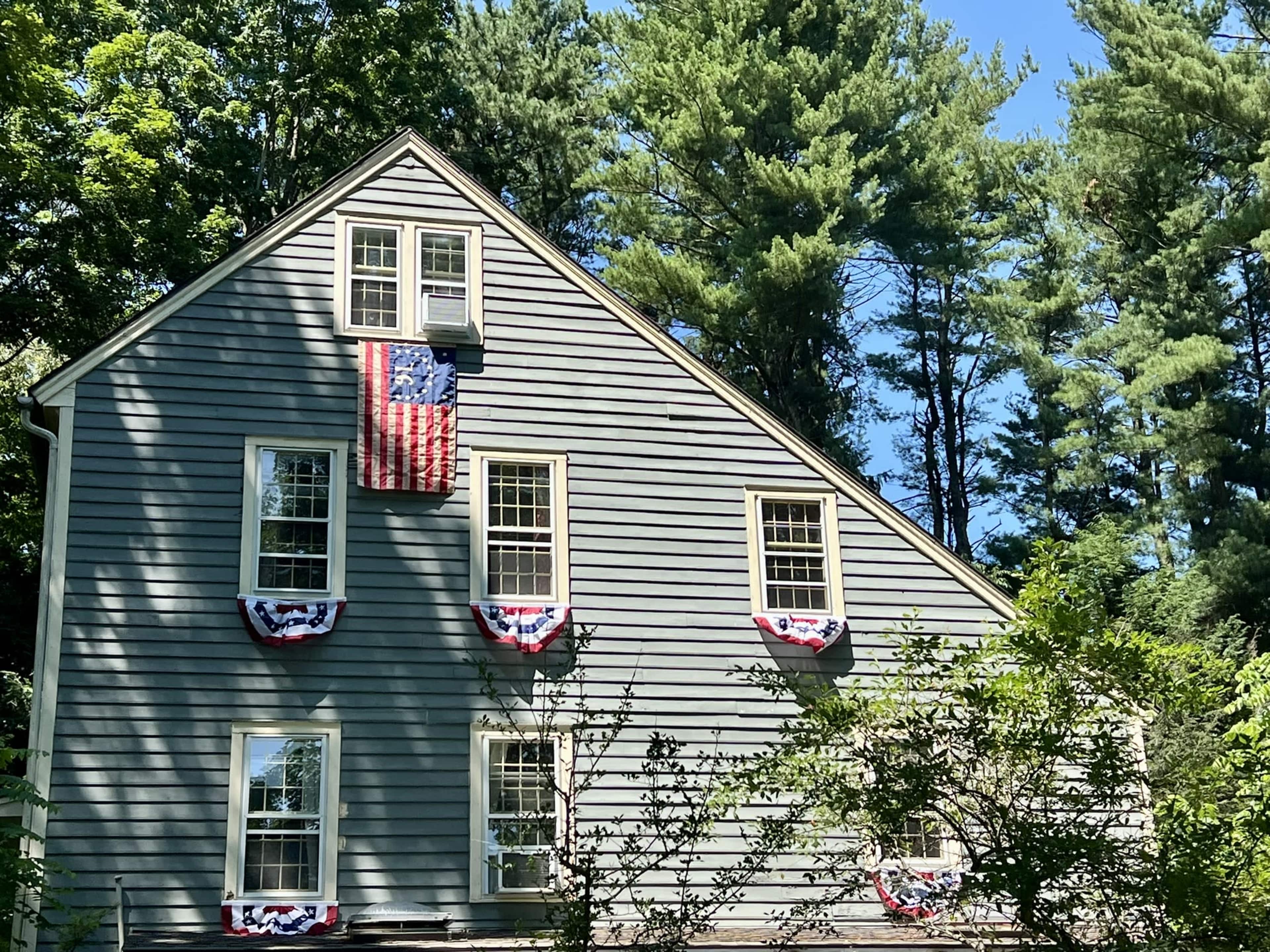 A gray house with multiple windows displays an American flag and red, white, and blue bunting under the windows, surrounded by tall trees.