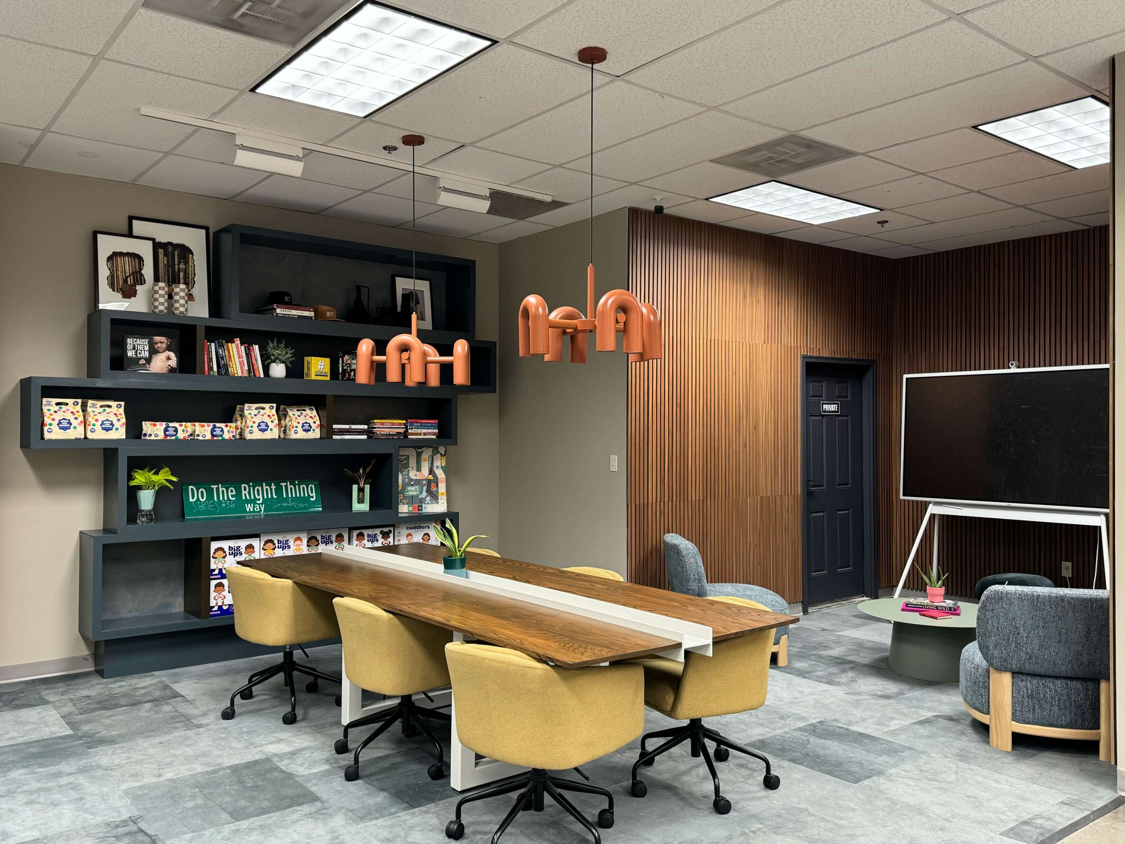 A modern meeting room featuring a long wooden table surrounded by yellow chairs, with a dark green bookshelf, decorative items, and unique pendant lights overhead.