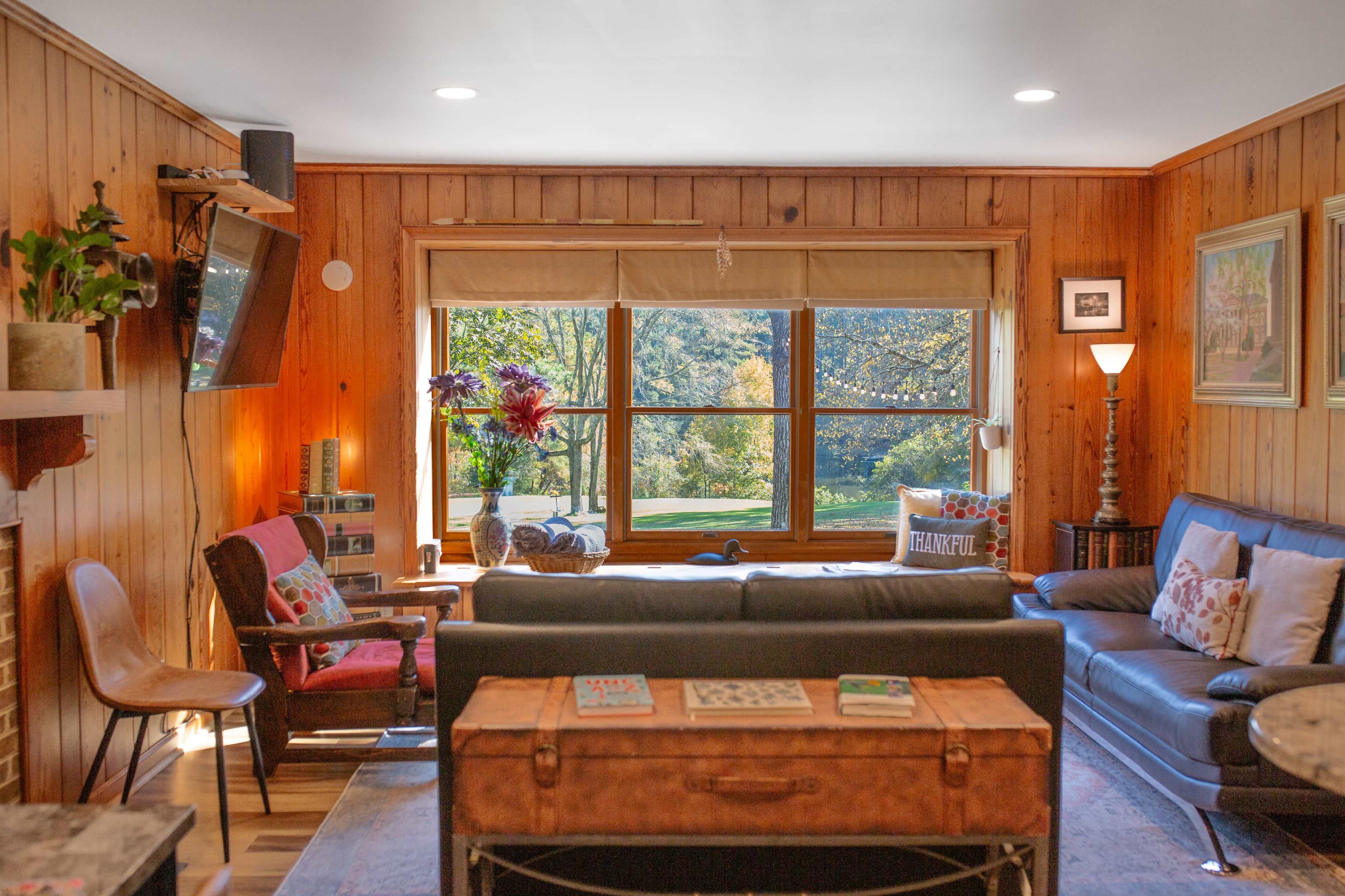 The image shows a cozy living room with wooden paneling, featuring a dark leather sofa, a tufted coffee table, and large windows overlooking a green landscape.