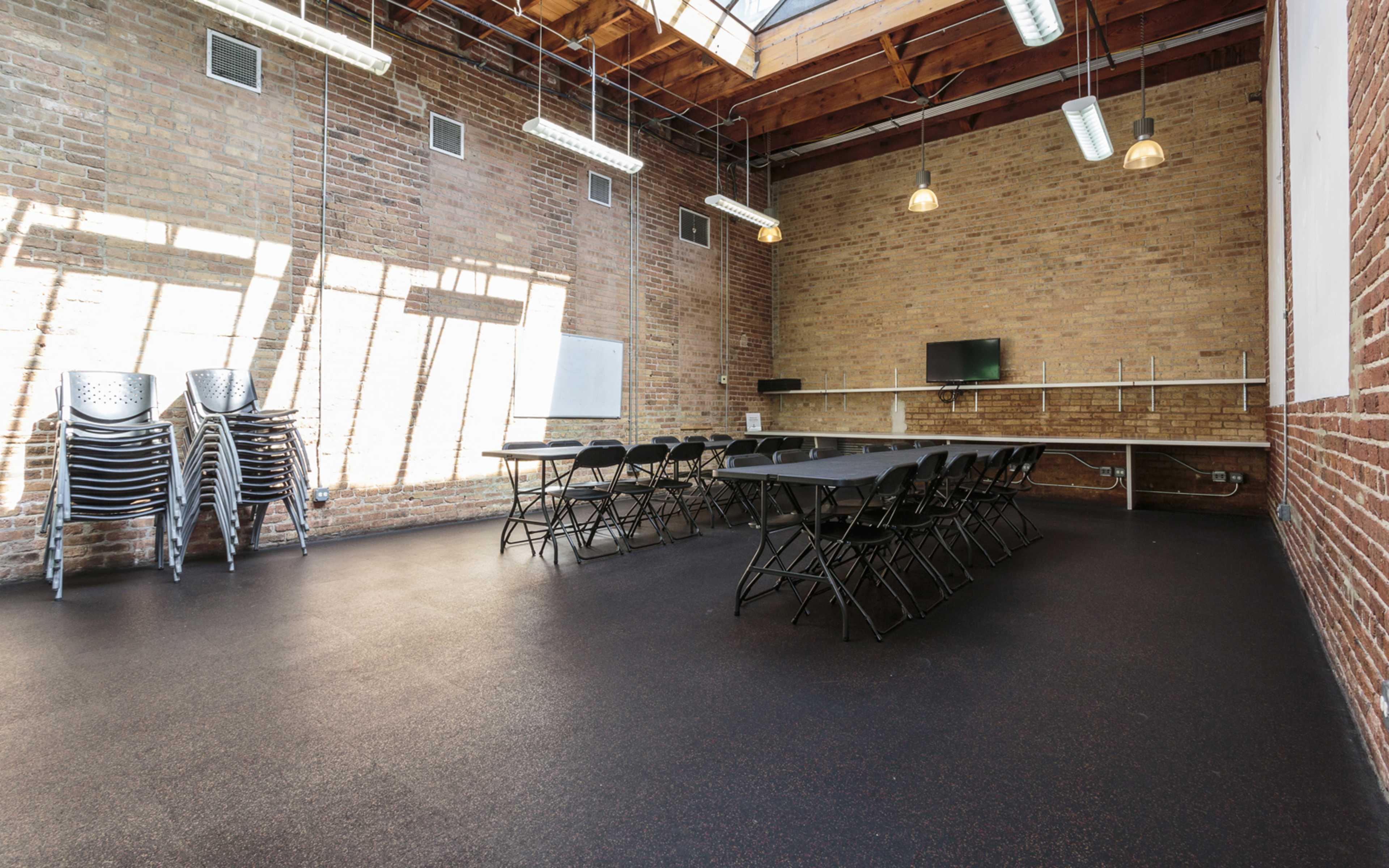 A spacious room with a long black table surrounded by chairs, large windows allowing natural light, and stacked metal folding chairs against a brick wall.