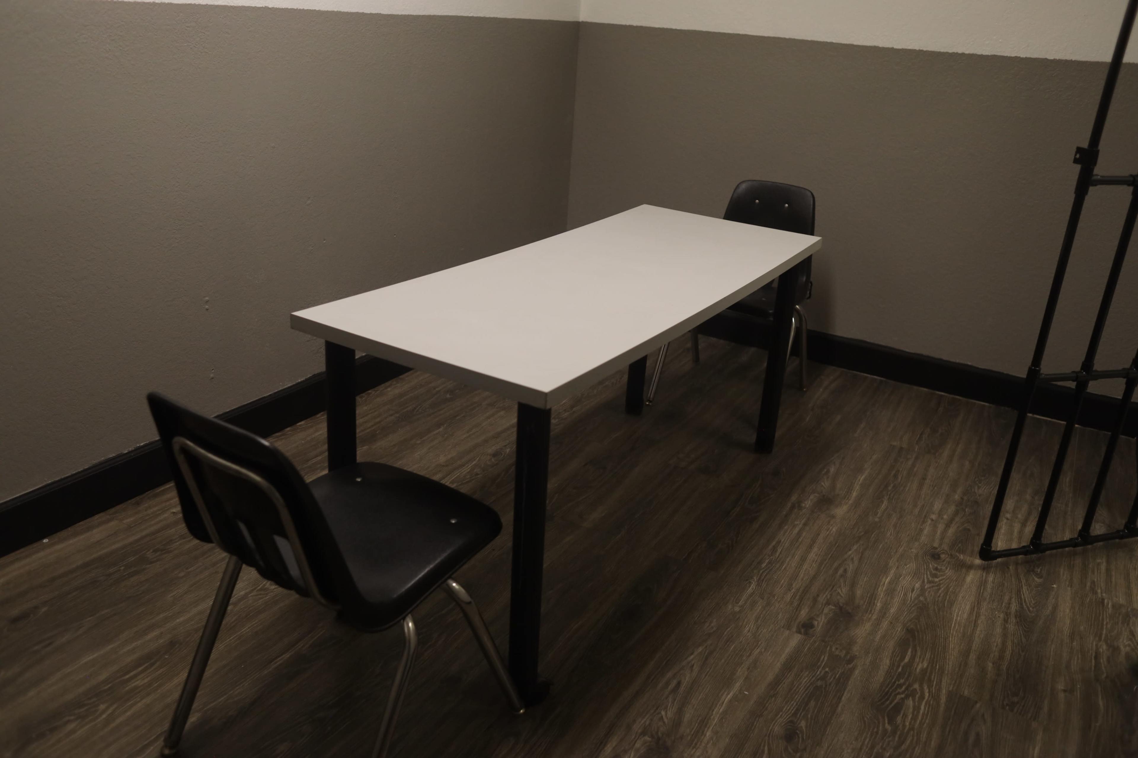 The image shows a small, sparsely furnished room with a white table and two black chairs on a wooden floor.