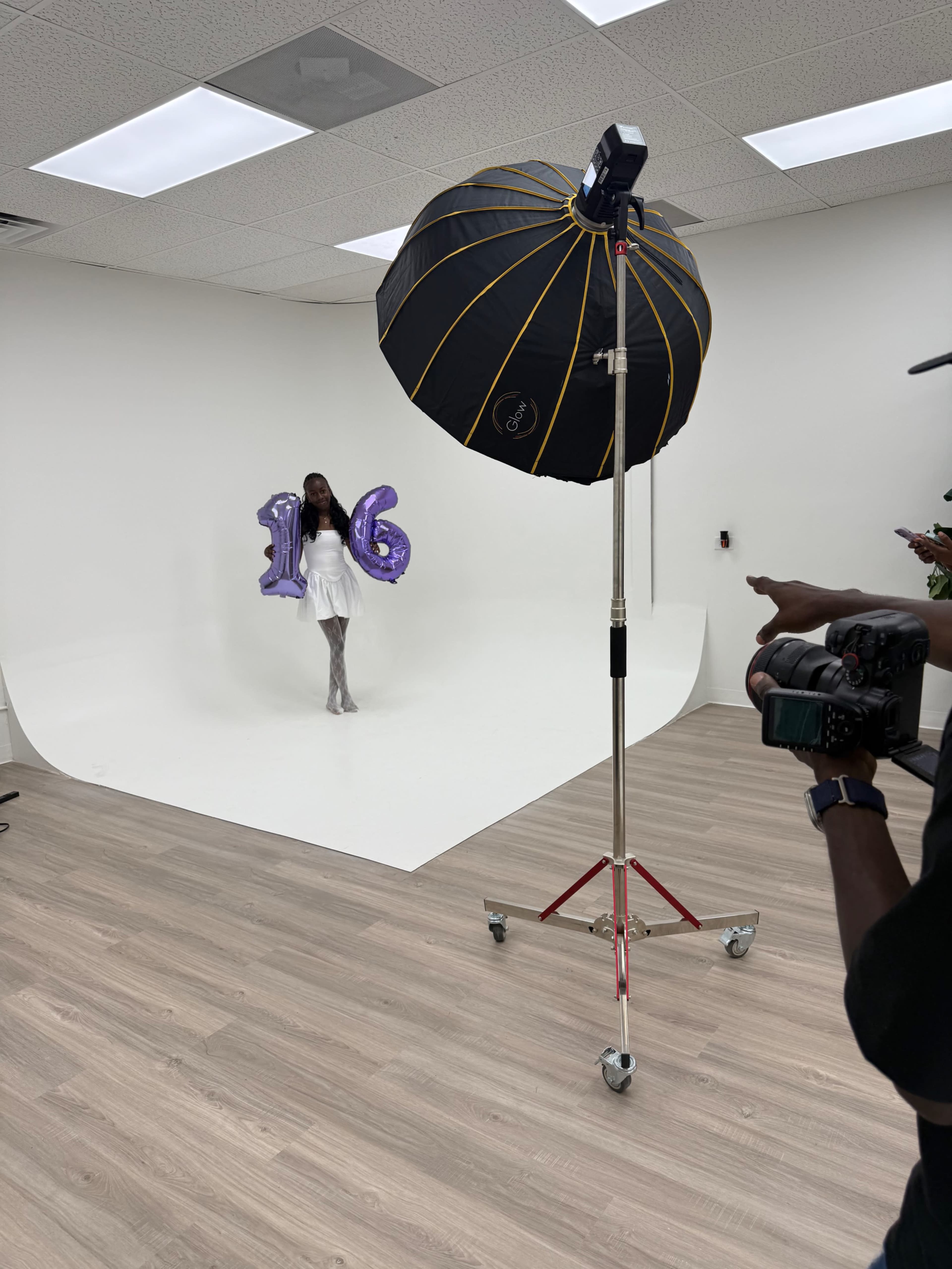 A model poses with large purple "16" balloons in a studio setting, illuminated by a softbox light.