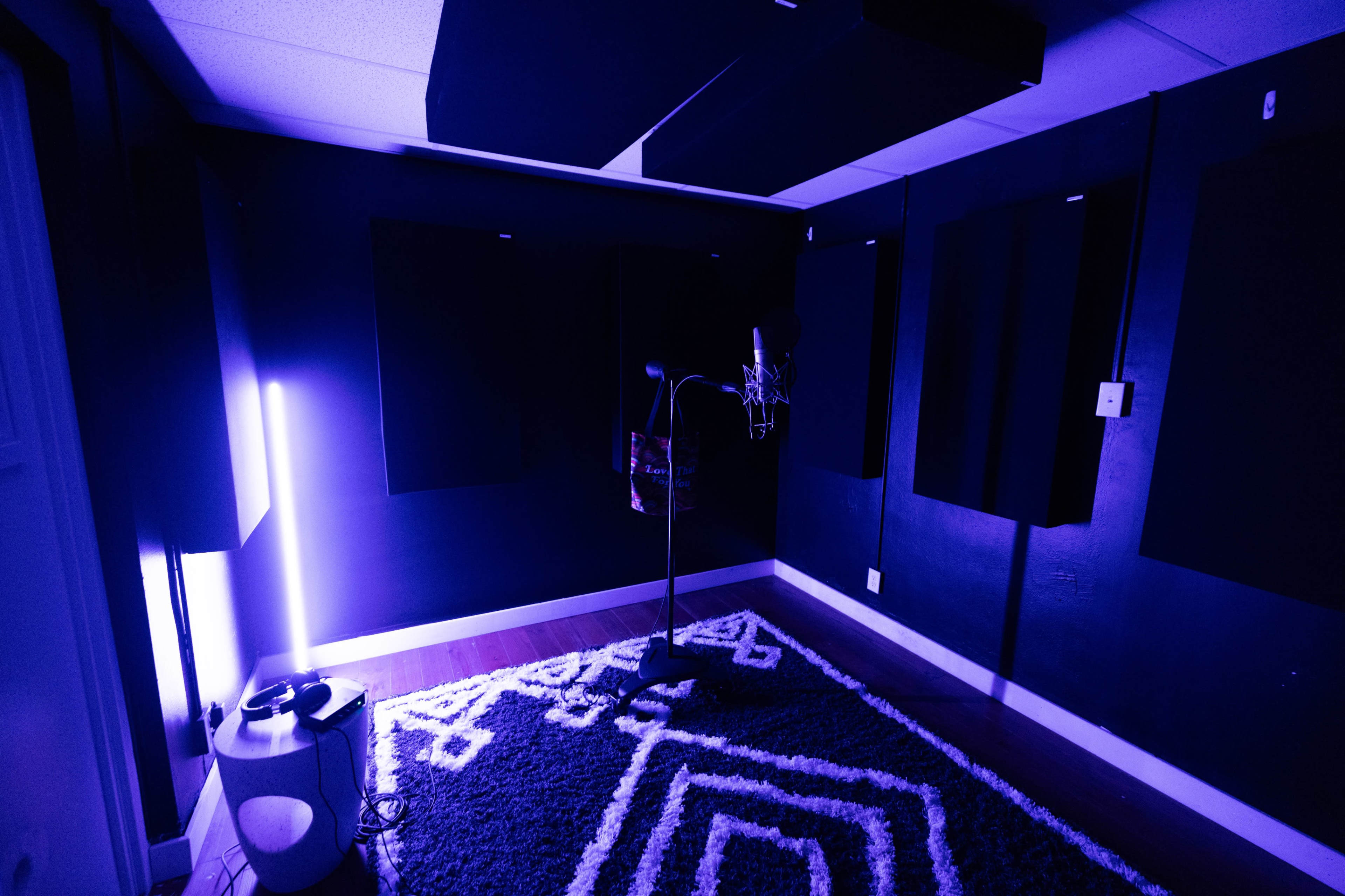 A small recording studio features a microphone on a stand, surrounded by soundproofing panels and illuminated by purple lighting.