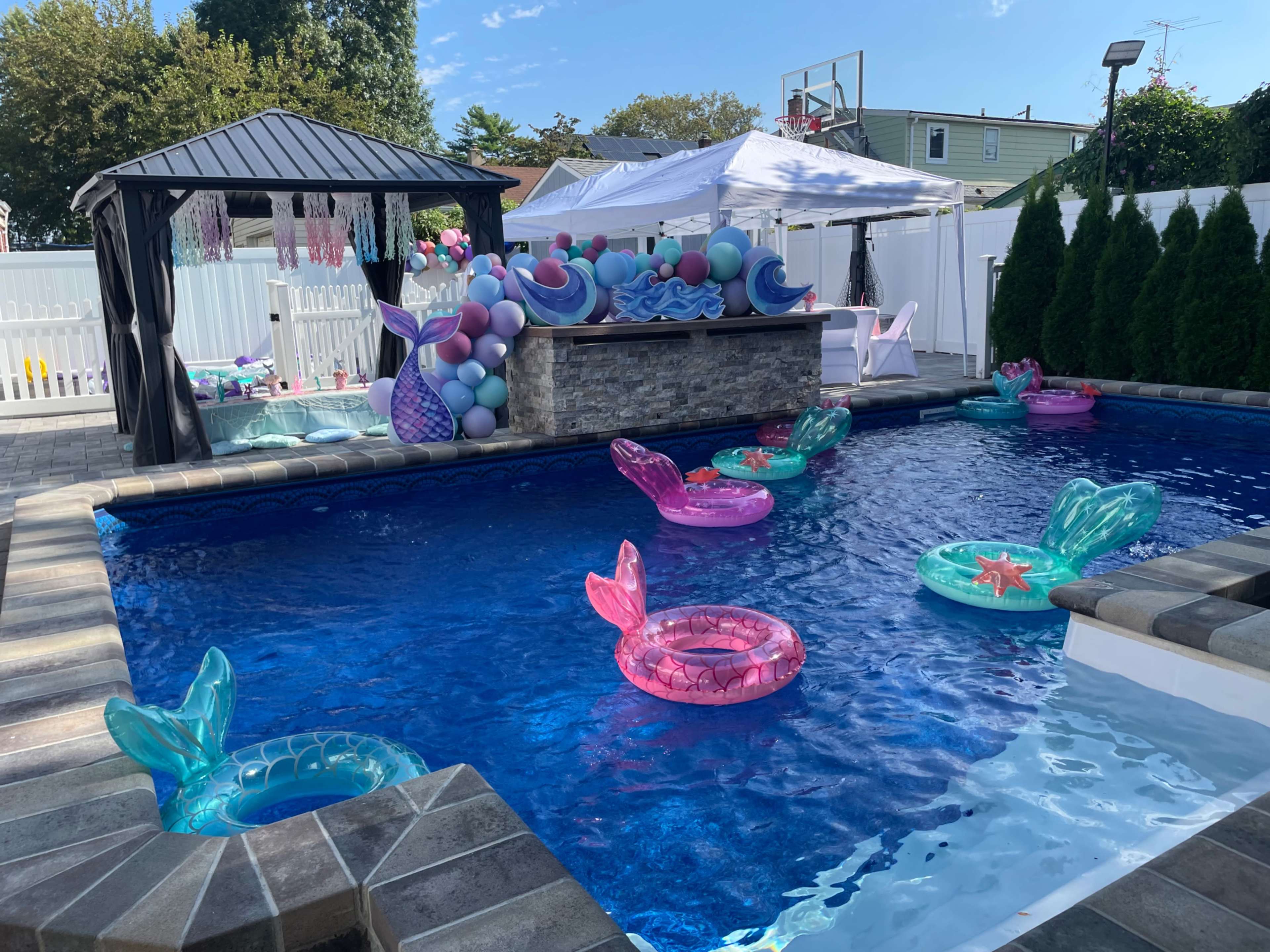 A backyard pool features colorful mermaid-themed floaties, a stone bar area, and decorative tents filled with balloons.