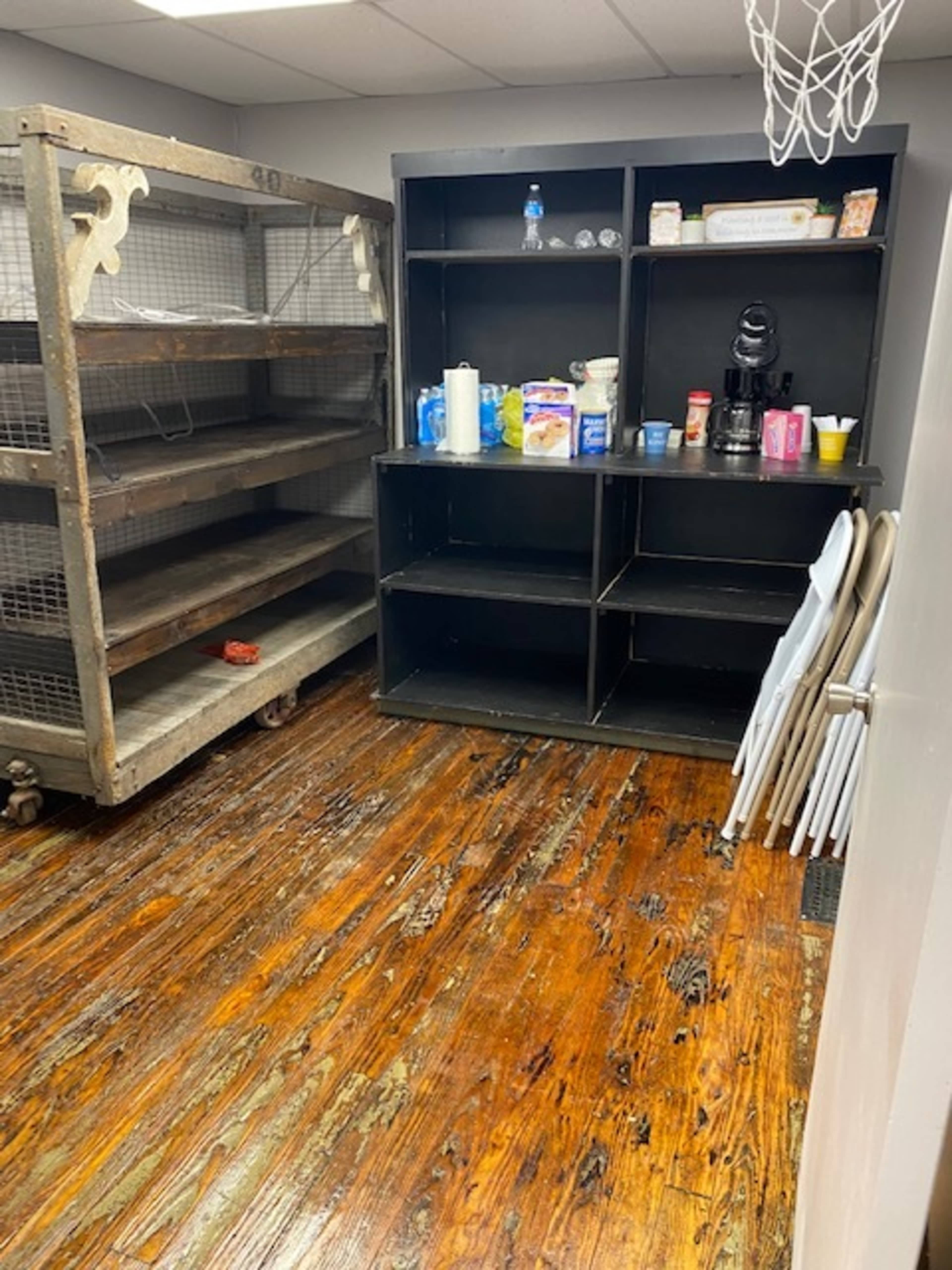 The image shows a room with wooden flooring, featuring a metal shelving unit on the left and a black shelving unit filled with various items on the right.