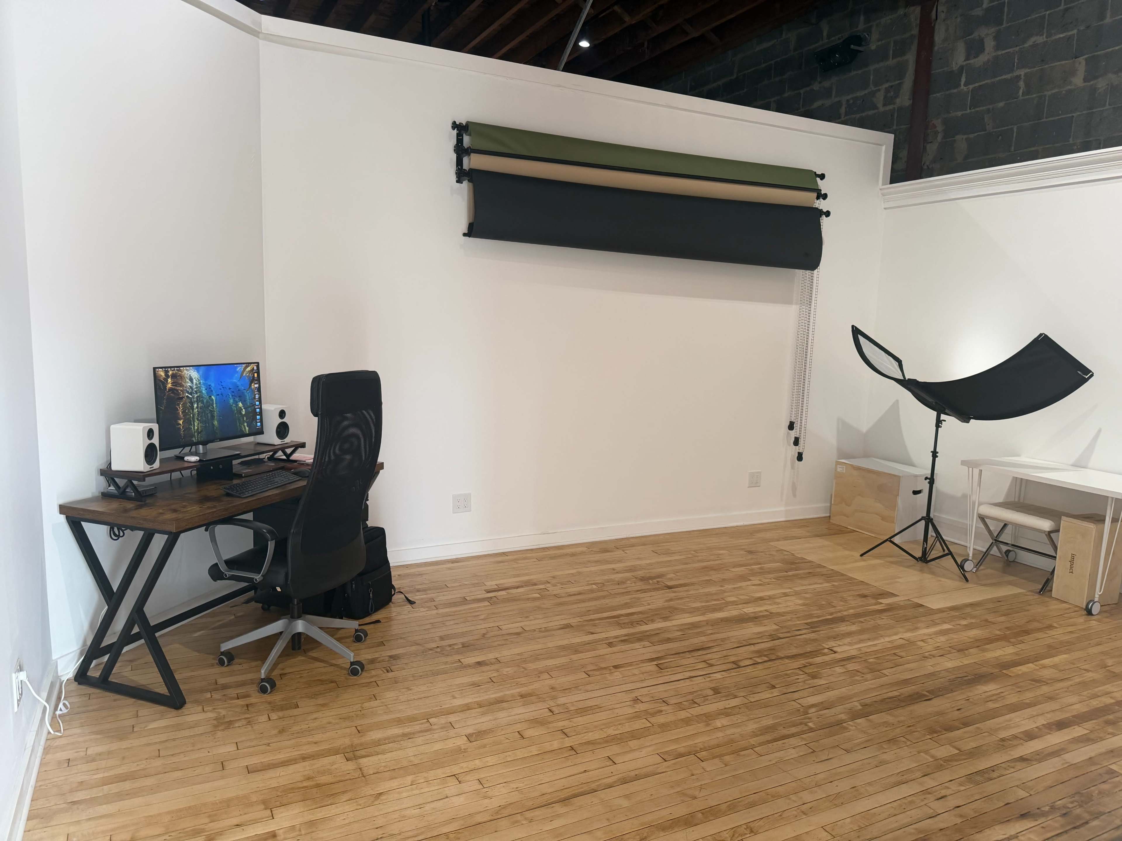 The image shows a minimalistic room featuring a wooden desk with a computer monitor, an ergonomic chair, a backdrop roller with green and black materials, and a photography light umbrella positioned in the corner.