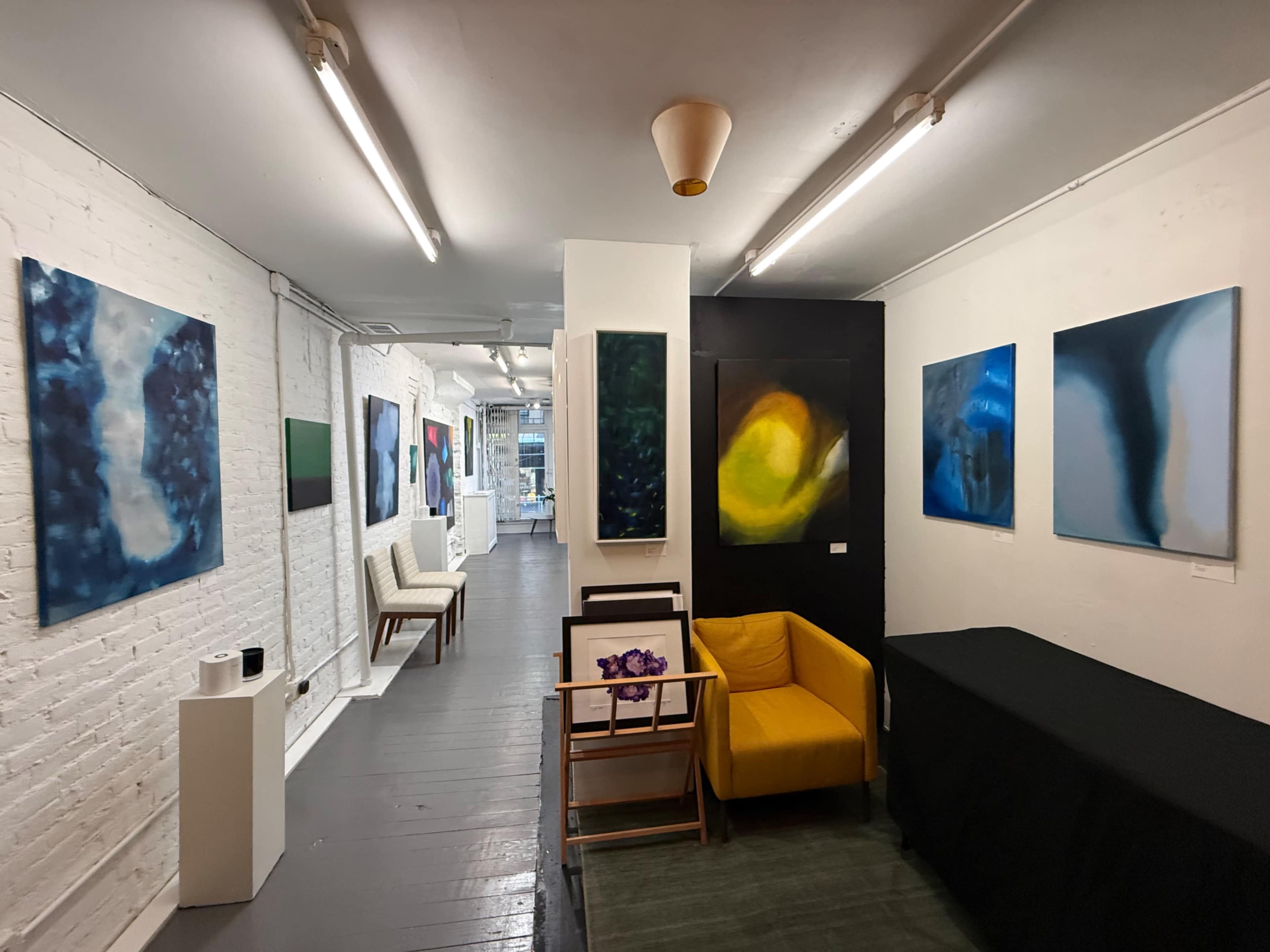 The image shows an art gallery featuring abstract paintings on white brick walls, with a yellow chair and a black table positioned in the foreground.