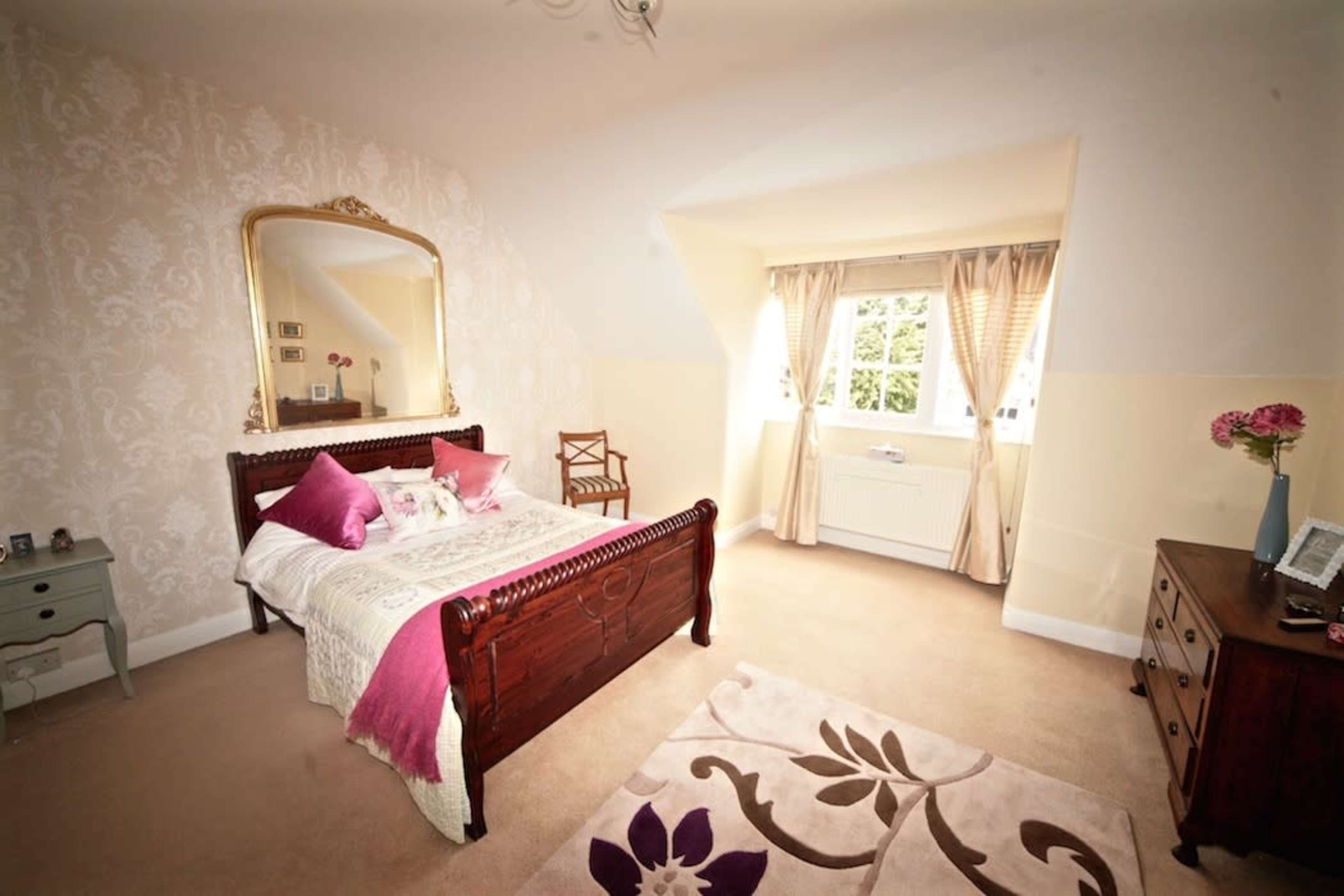 A cozy bedroom featuring a wooden bed with decorative pillows, a large mirror, a small chair, and a window with curtains.