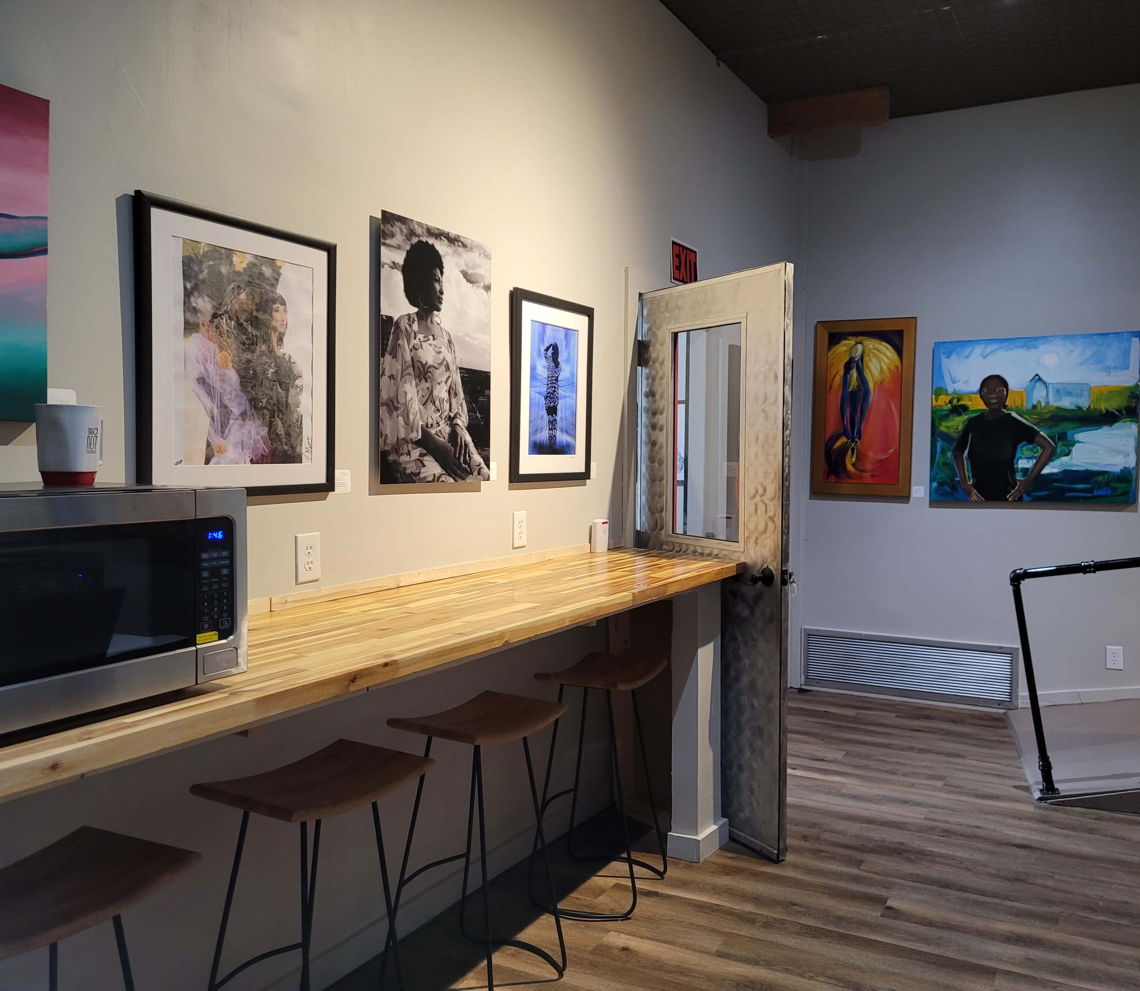 A cozy café interior featuring a wooden counter with stools, a microwave, and several framed artworks on the walls.