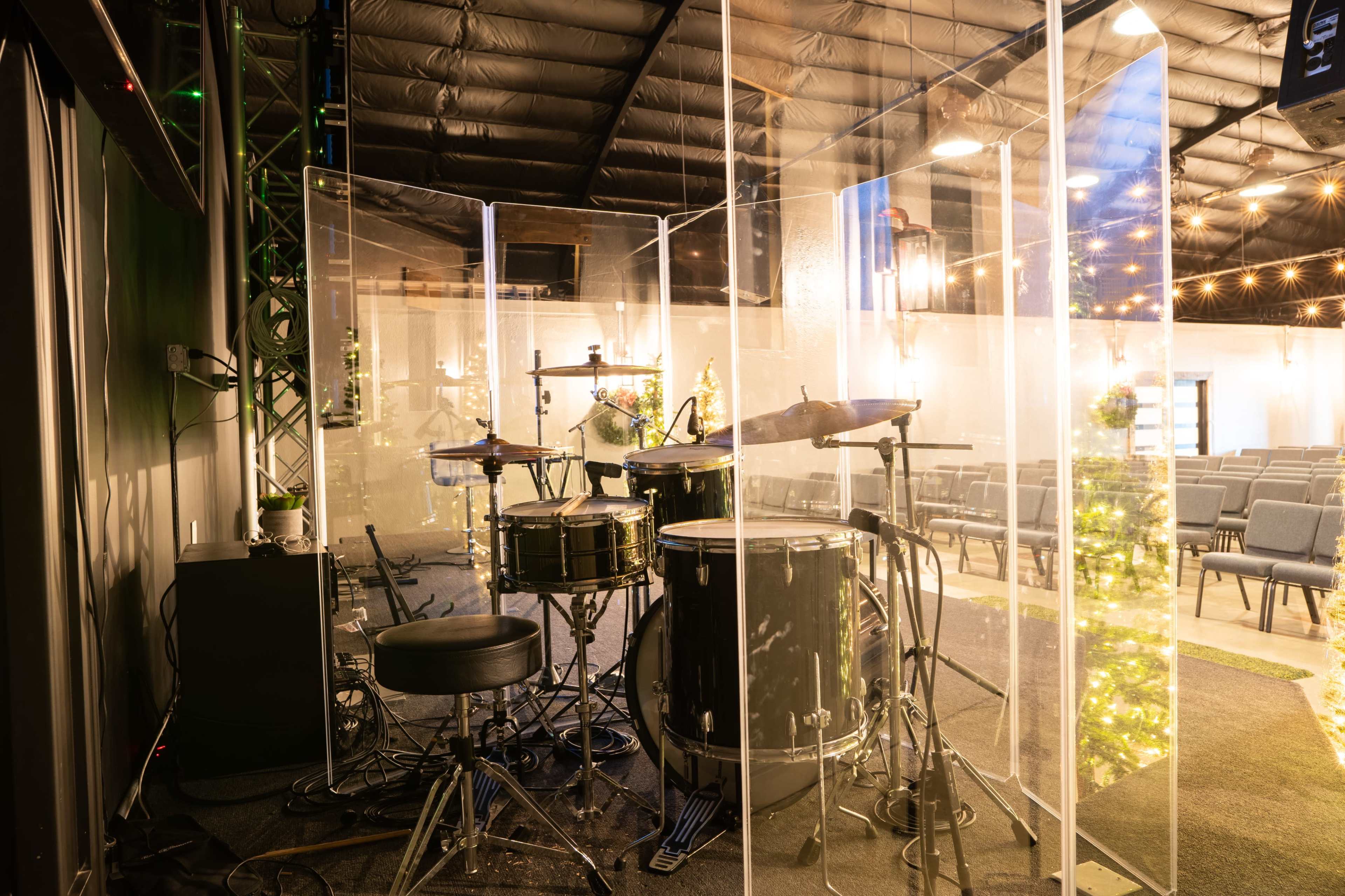 A drum set is positioned behind a clear acoustic shield in a spacious, empty auditorium with rows of chairs.