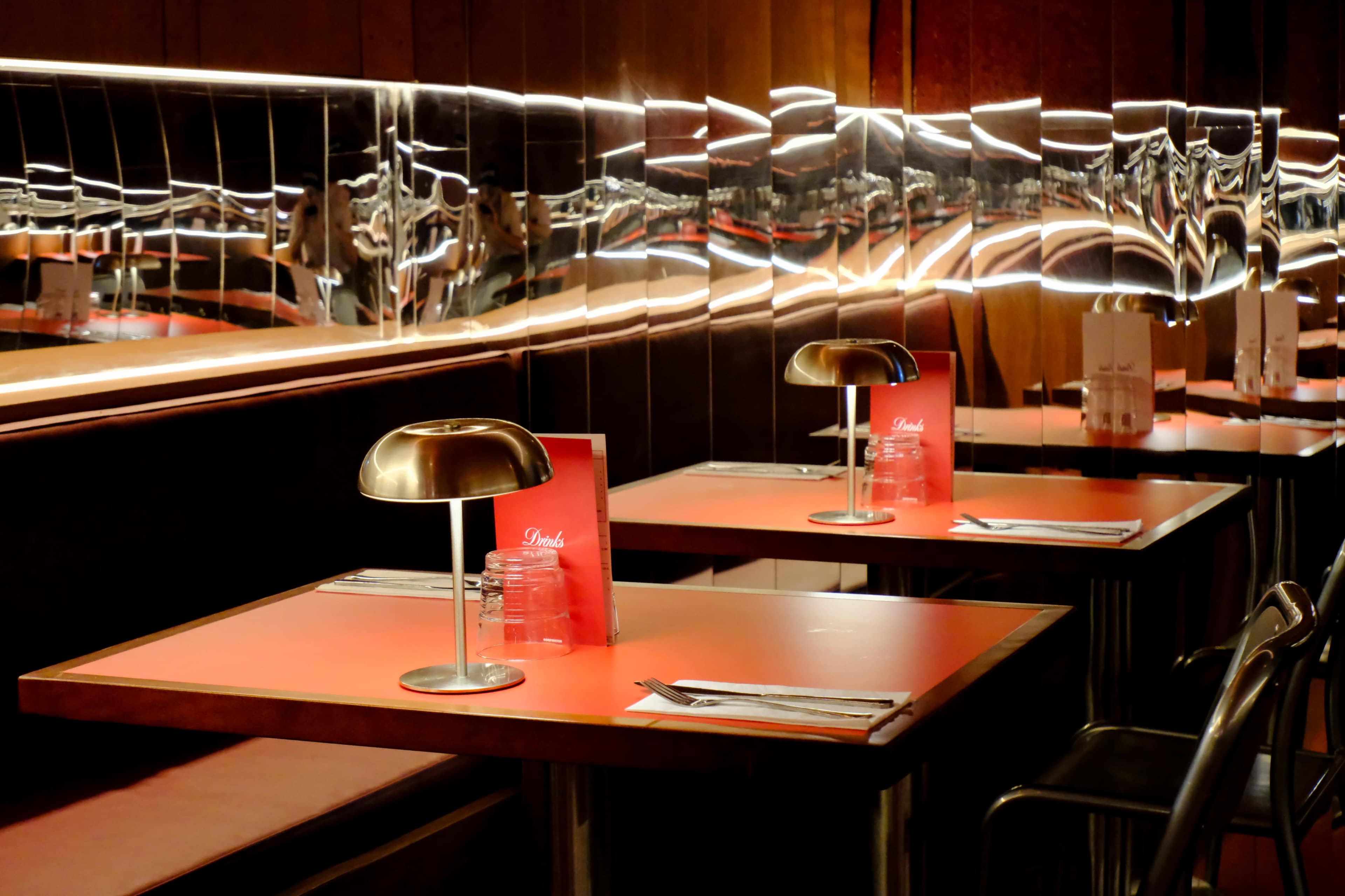 The image shows a modern restaurant interior with red tables, sleek metal lamps, and mirrored walls reflecting the surroundings.