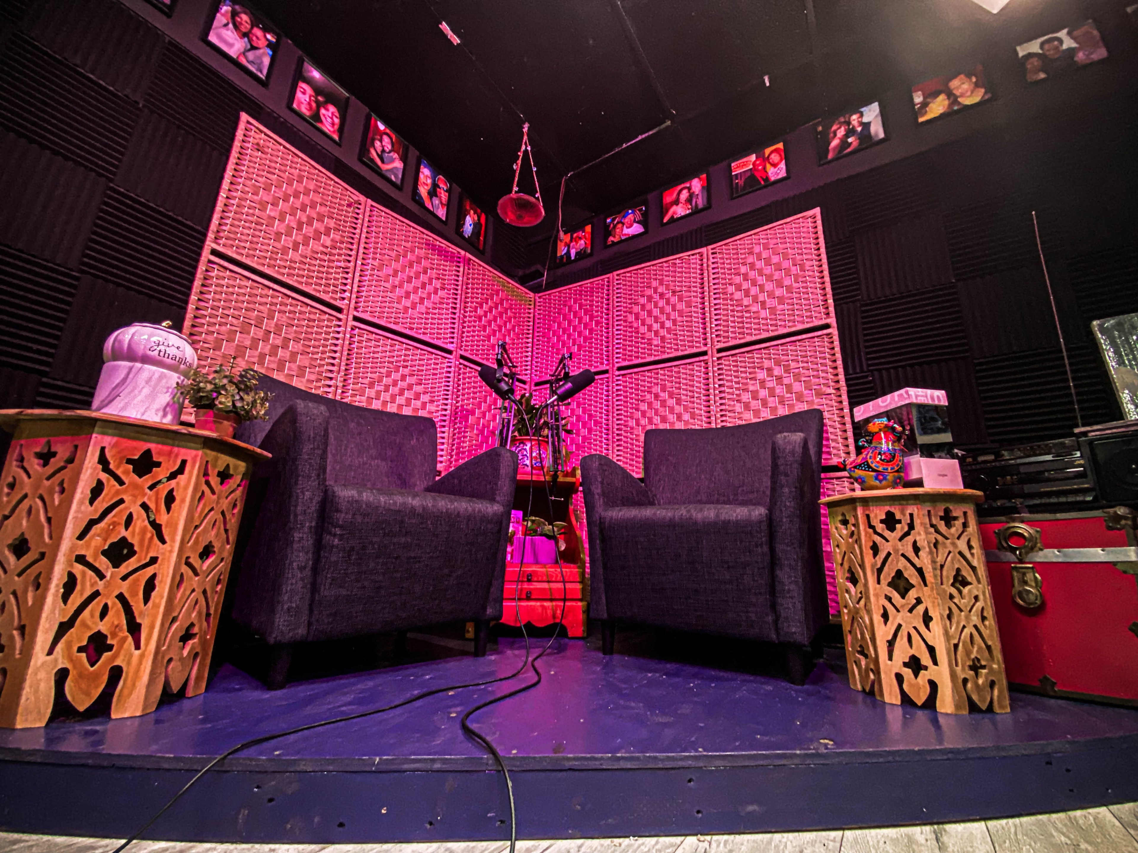 The image shows a colorful broadcasting or recording studio with two dark armchairs facing a microphone setup surrounded by patterned acoustic panels and framed photos on the walls.