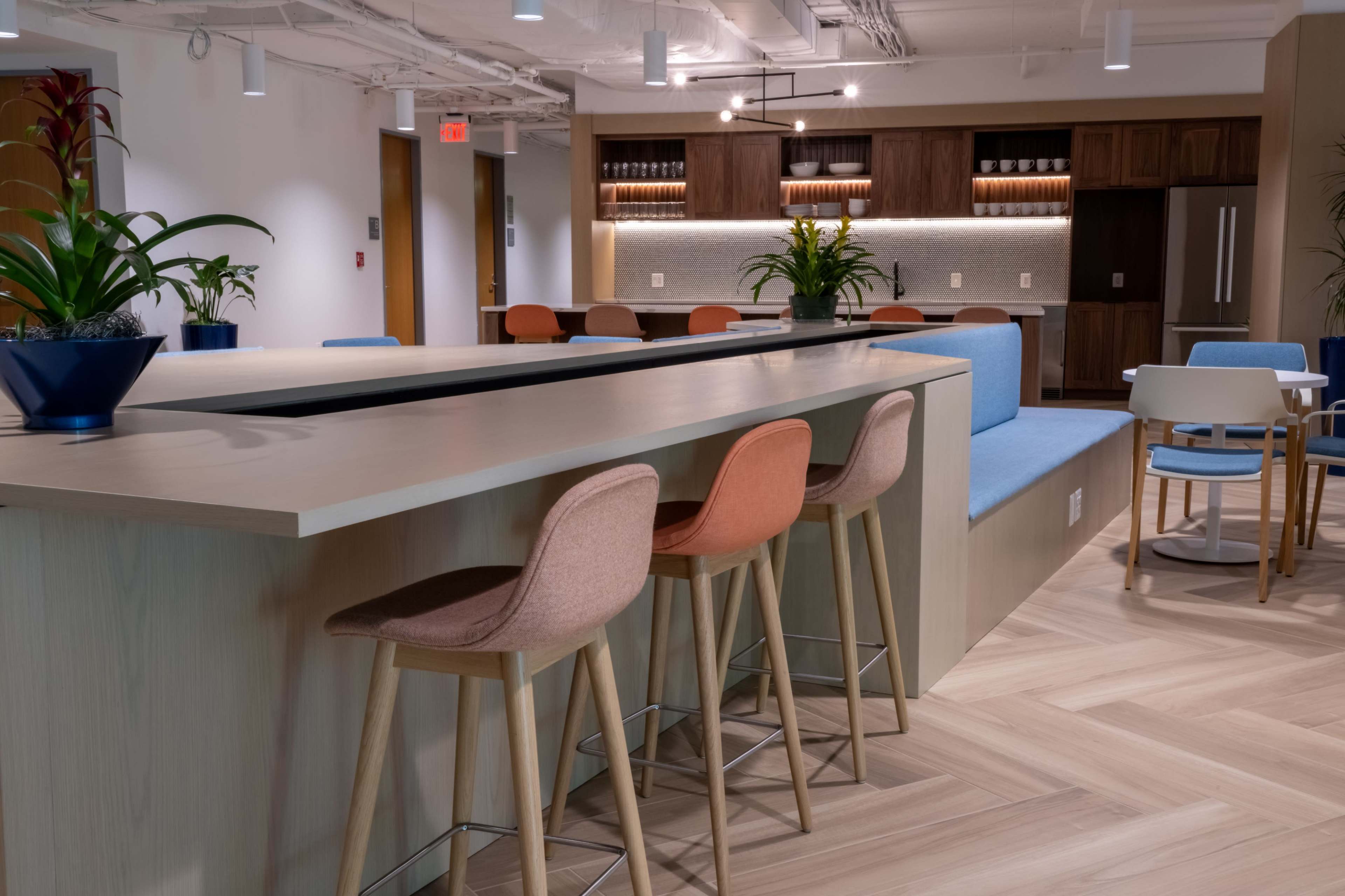 A modern communal kitchen area with a long wooden counter, several stools, and a seating nook against the wall.