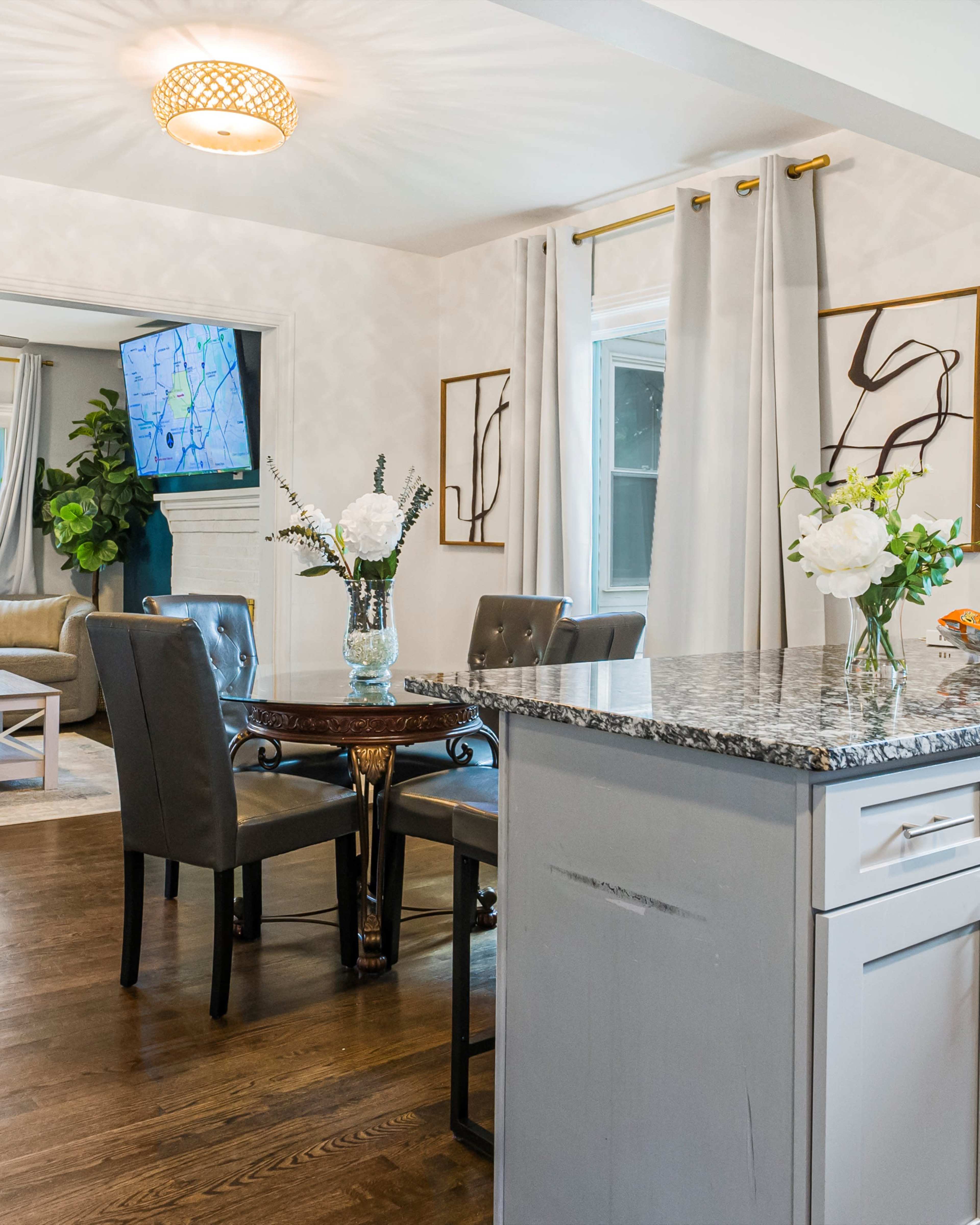 A dining area features a round table with four upholstered chairs, surrounded by light-colored walls, decorative art, and a nearby TV, with a kitchen island in the foreground.