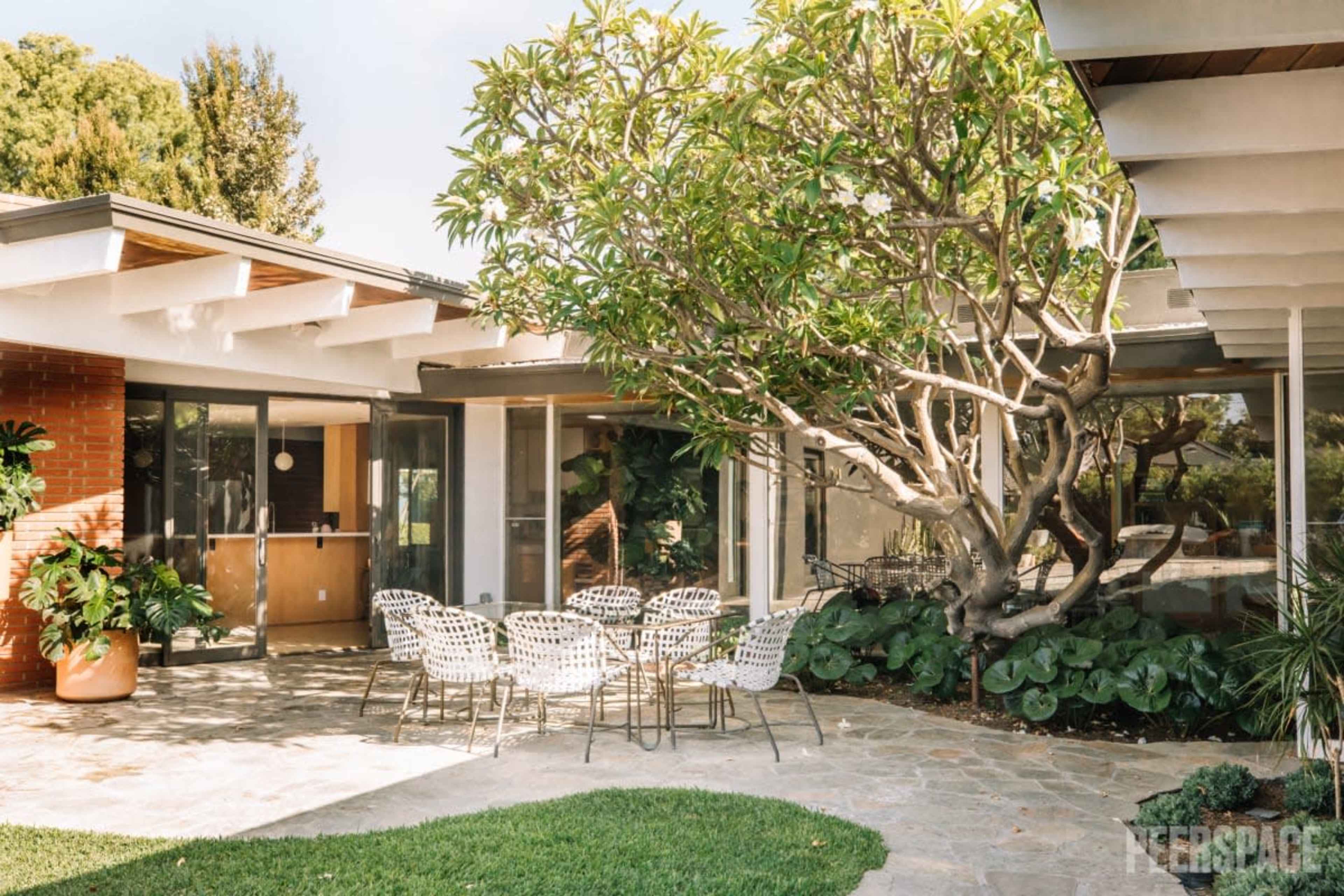 A modern patio features a round table surrounded by chairs, set in front of a house with large glass windows and a tree nearby.