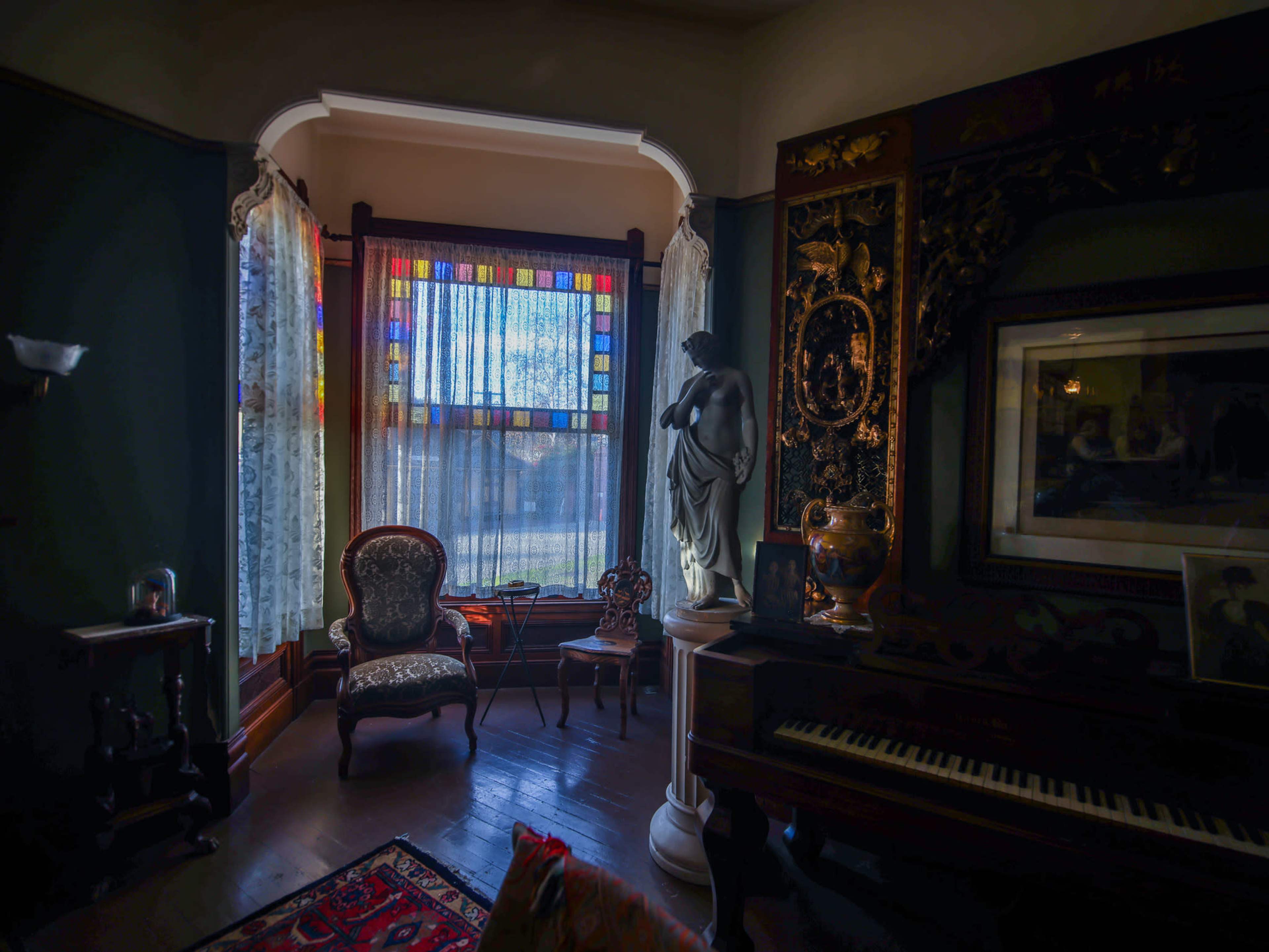 A vintage living room with a stained glass window, ornate furnishings, a statue, and a piano.