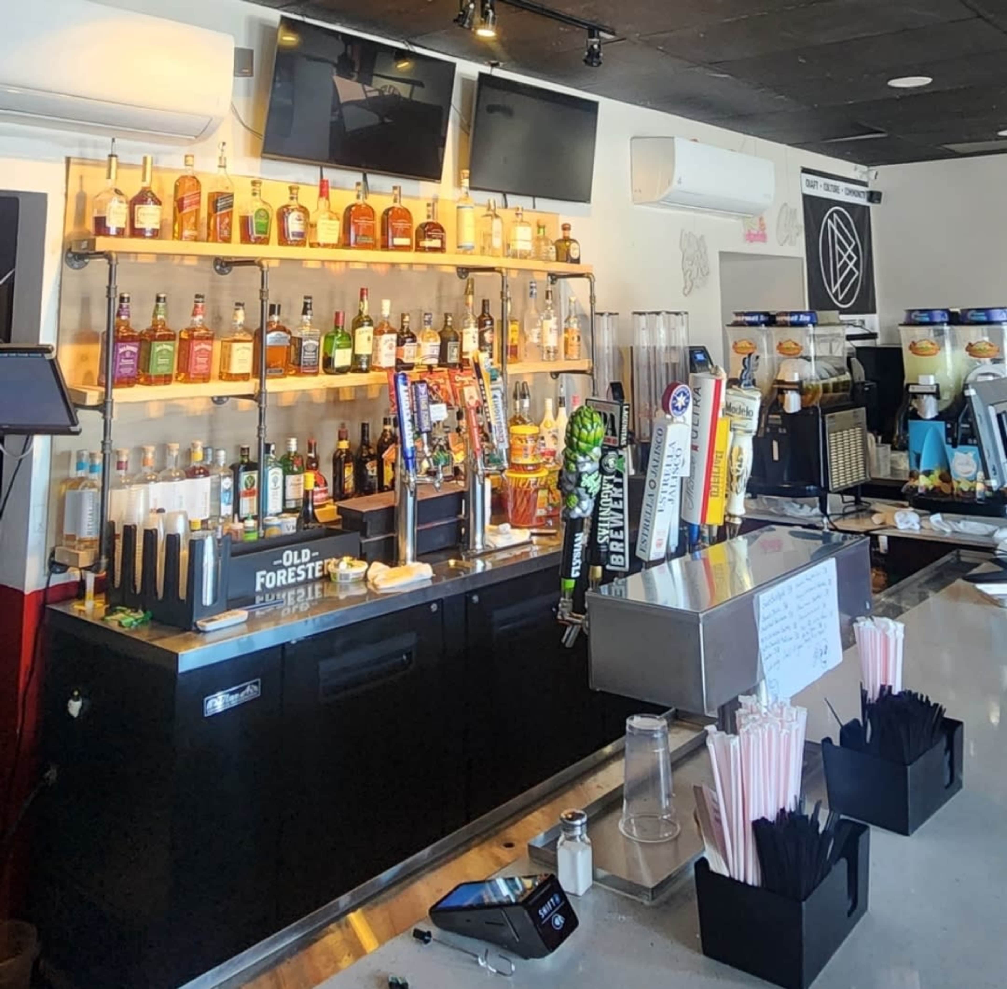 The image shows a bar area with a variety of liquor bottles displayed on shelves and several beer taps, alongside a counter with bar tools and straw dispensers.